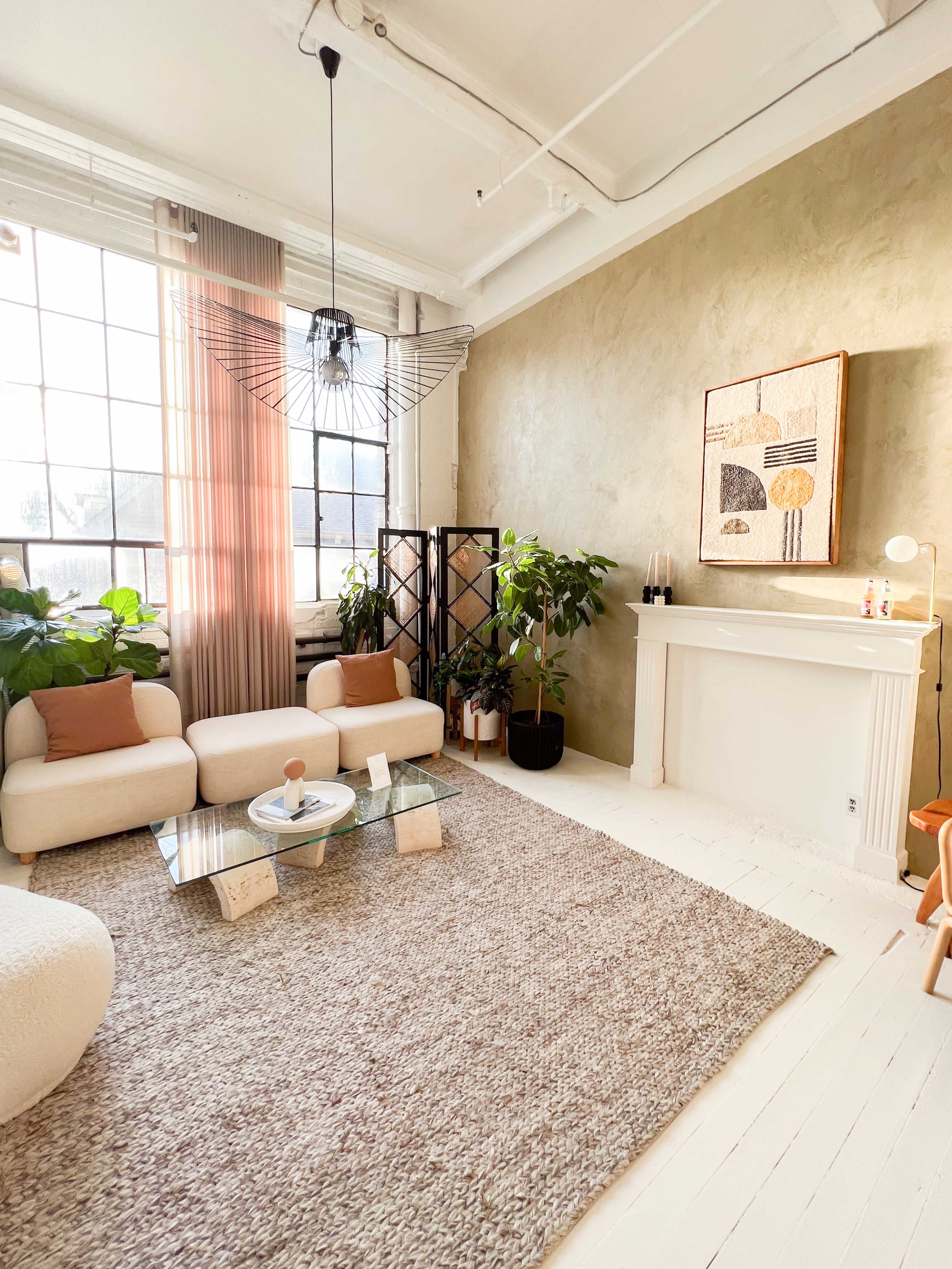 A well-lit living room featuring a textured green wall, a large window, a cozy seating area with white chairs, potted plants, and a decorative rug.
