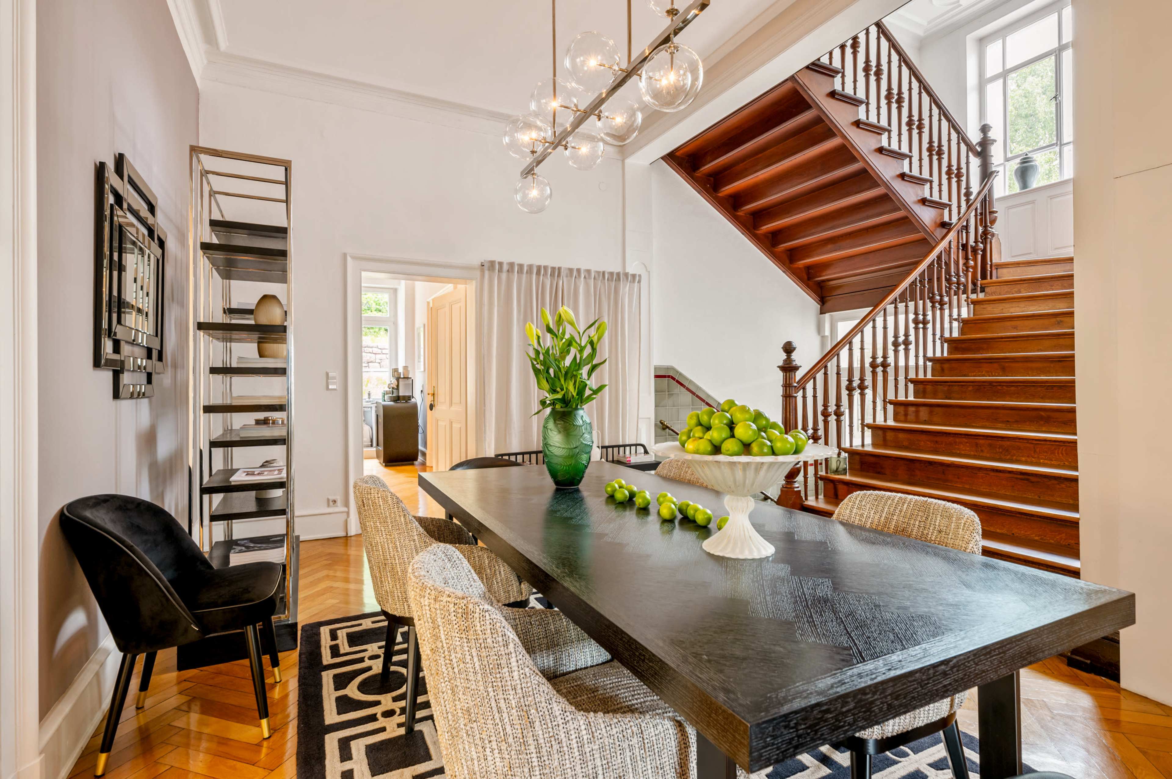A dining room features a black table surrounded by textured chairs, a bowl of limes, and a vase of tulips, with a staircase in the background.