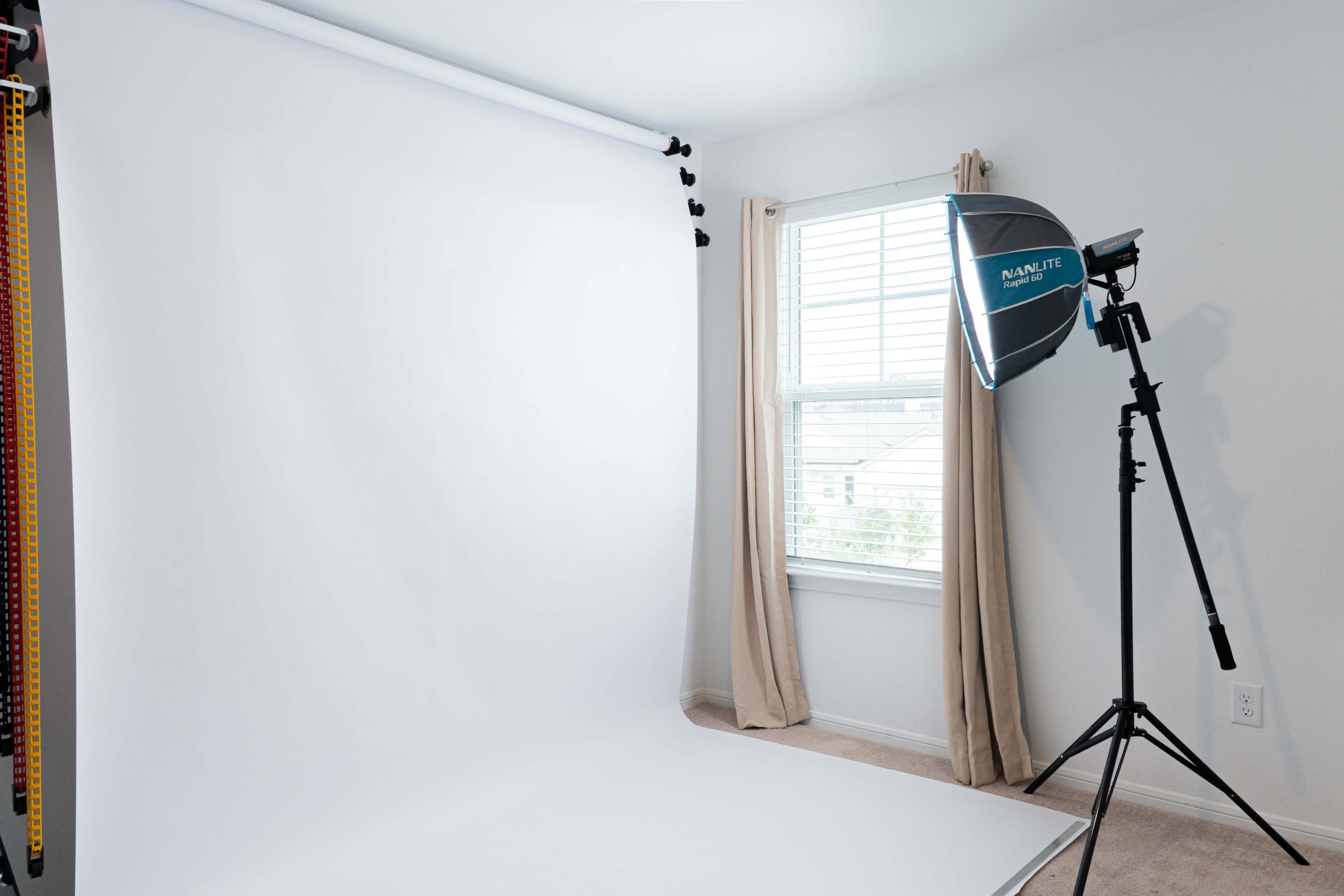 The image shows a photography setup with a white backdrop and a lighting kit positioned beside a window in a room.