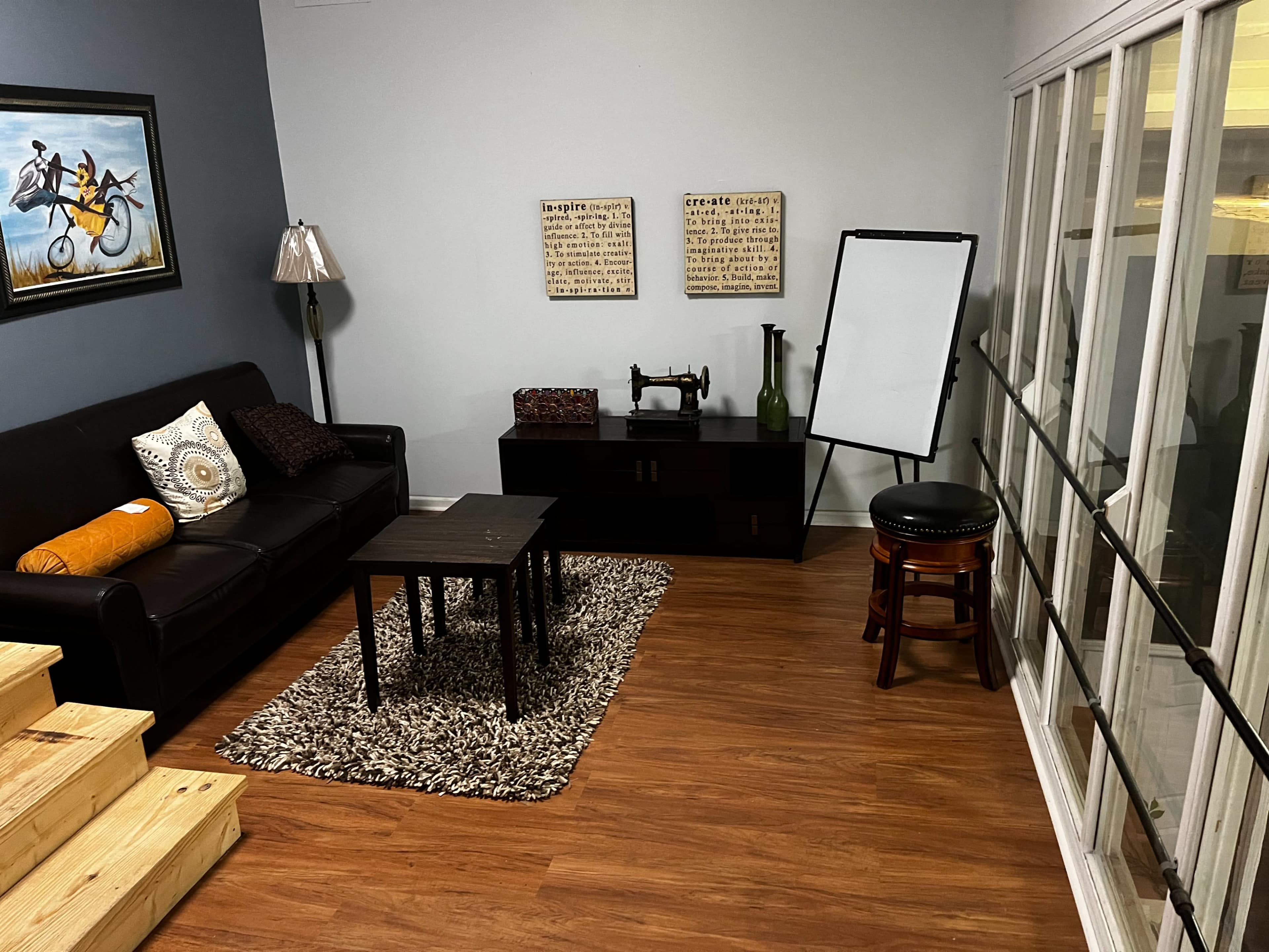 The room features a black sofa, a coffee table, decorative wall art, and a standing whiteboard in a space with wooden flooring and steps leading to it.
