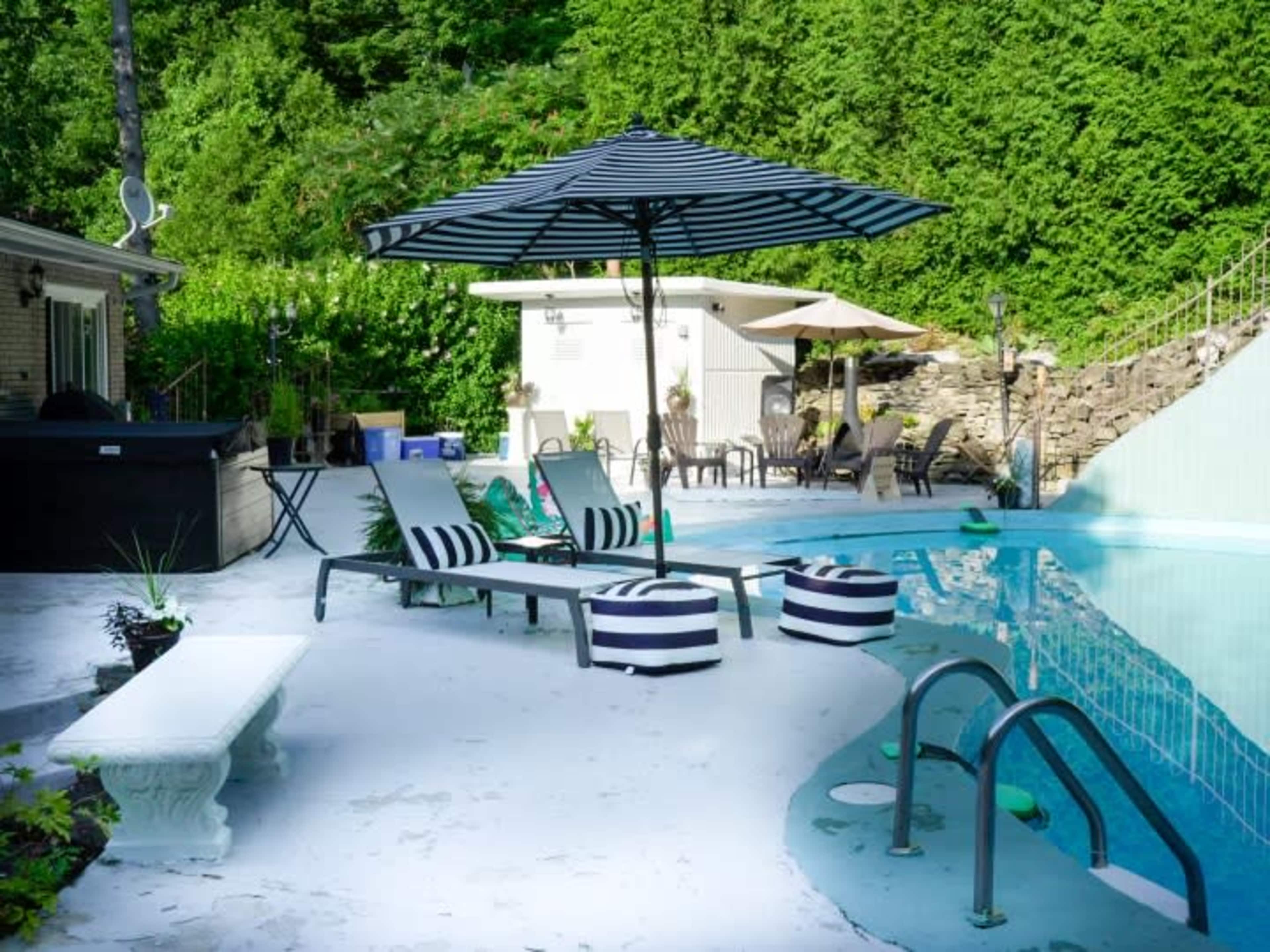 A swimming pool area features several loungers, umbrellas, and a concrete deck surrounded by greenery.