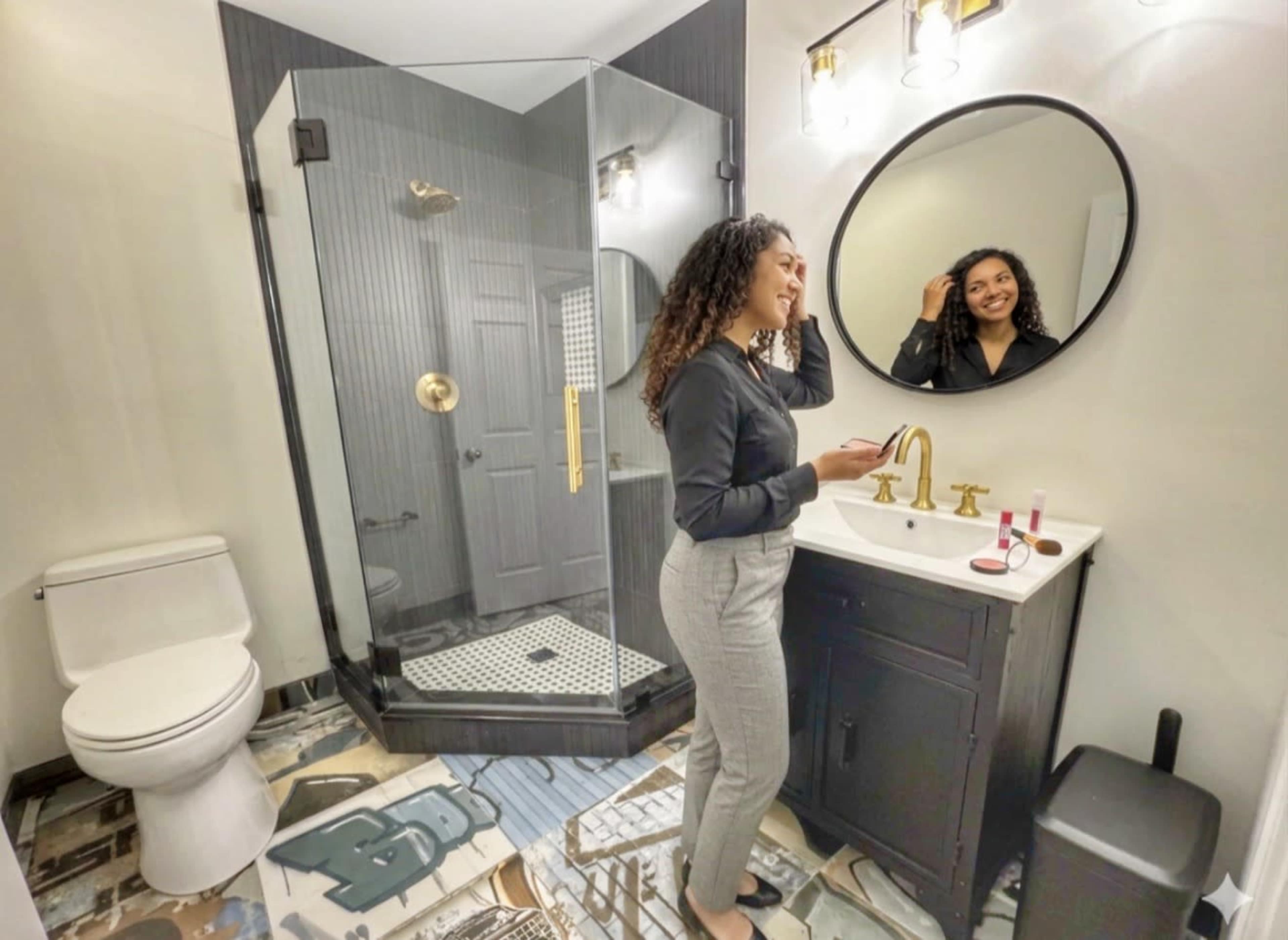 A woman stands in a modern bathroom with a glass shower, a round mirror above the sink, and a decorative floor.
