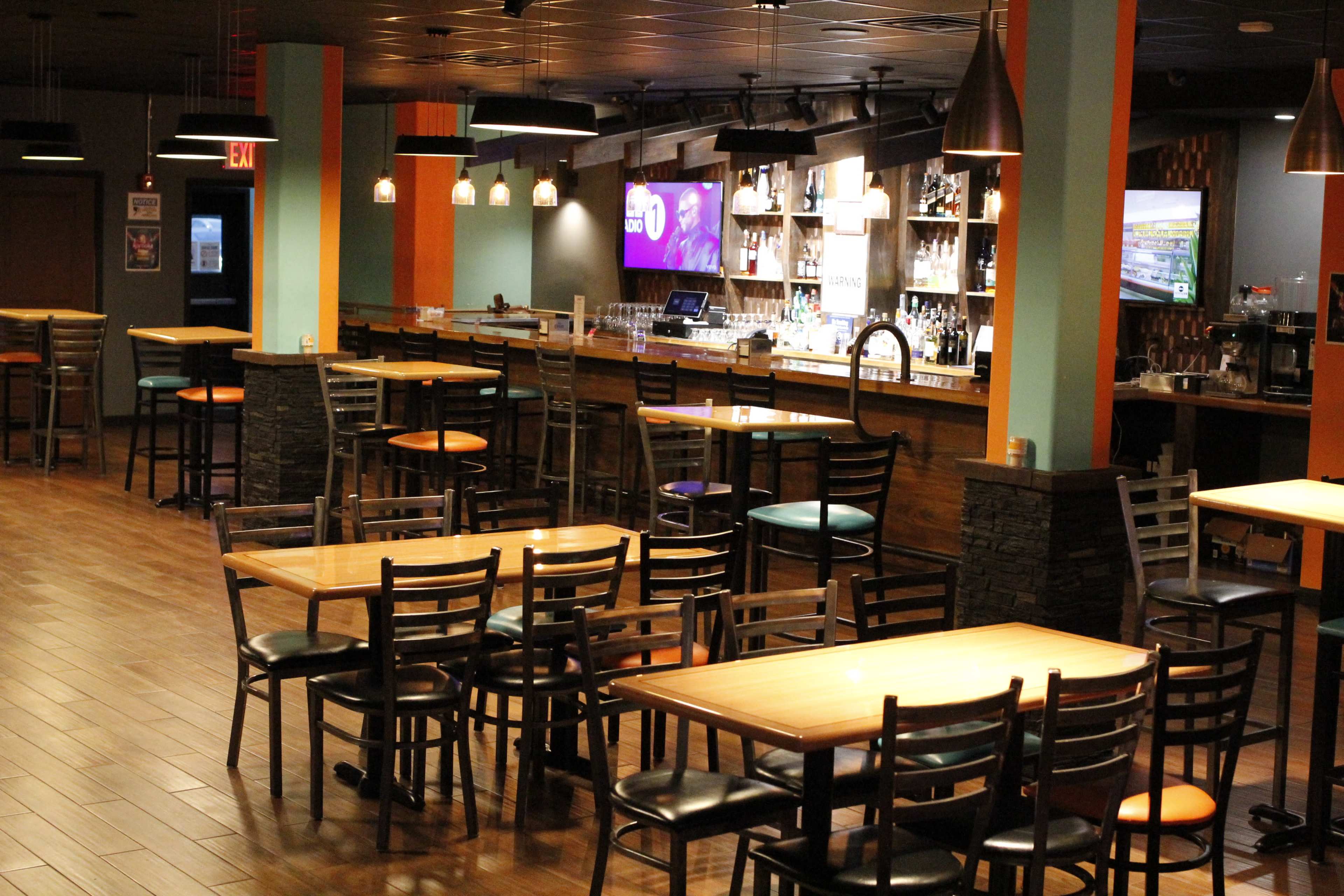 A dimly lit bar and dining area features several wooden tables and chairs arranged throughout, with a long bar in the background displaying bottles and a television screen.