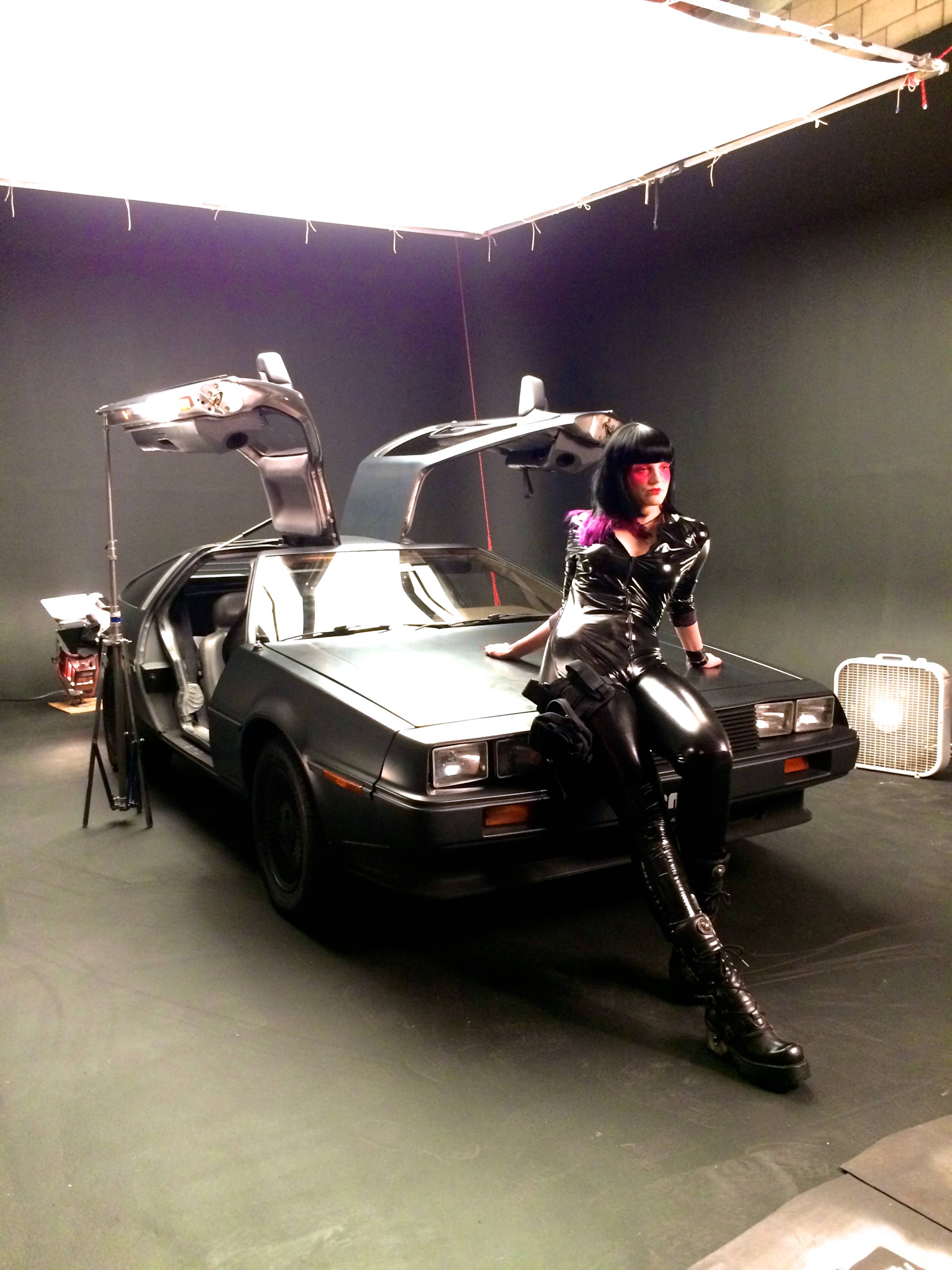 A person in a black outfit sits on the hood of a DeLorean car with its gullwing doors open in a studio setting.