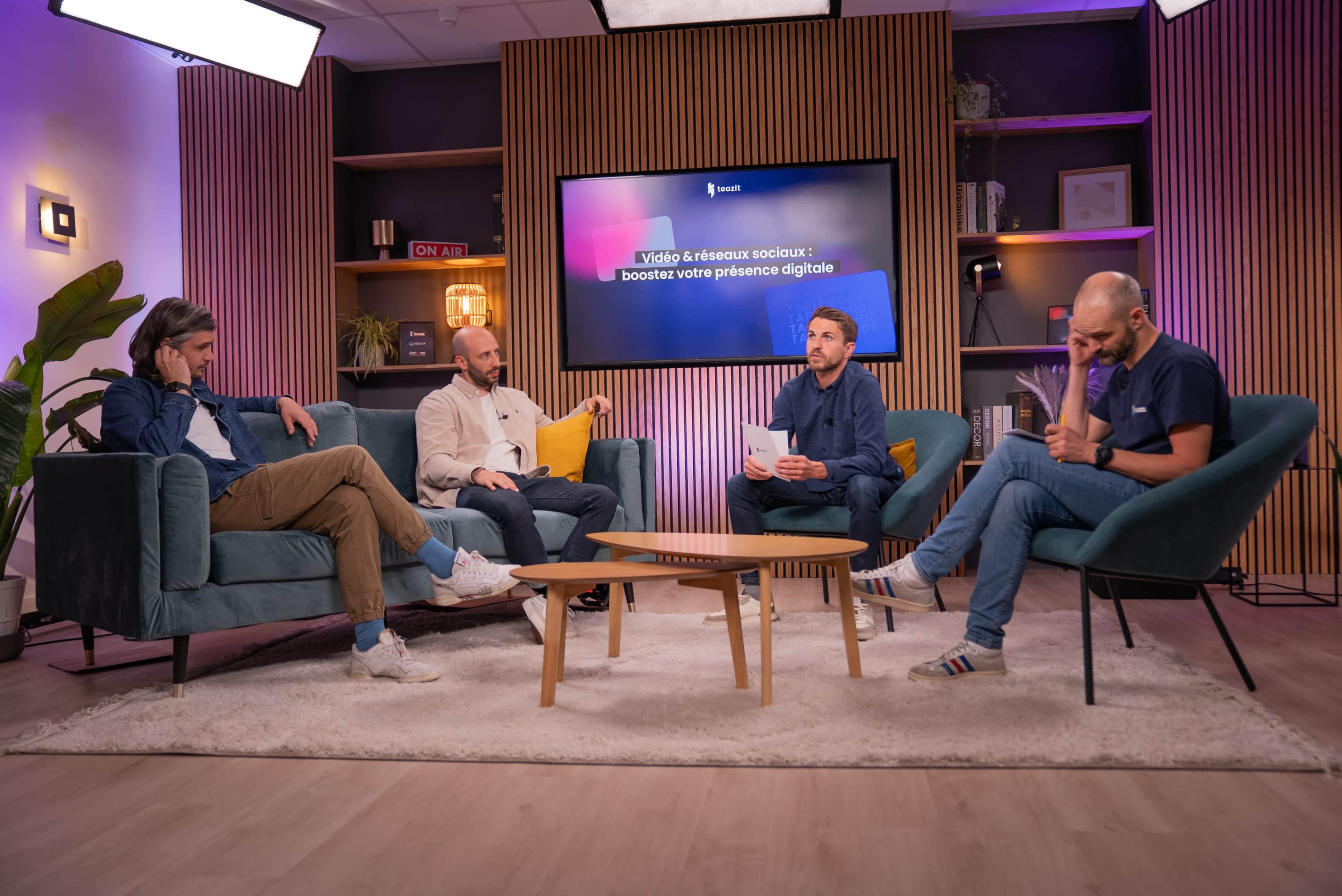 Four men are seated in a modern, well-lit room, engaging in discussion while various colorful screens are displayed in the background.