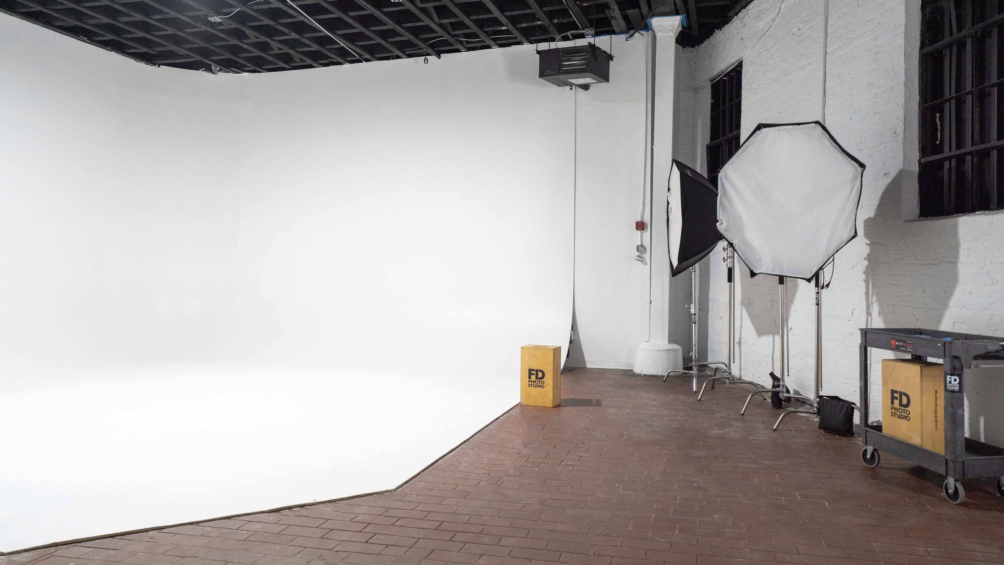 A photography studio featuring a white backdrop, a silver reflector, and a wooden storage crate in a brick-floored space.