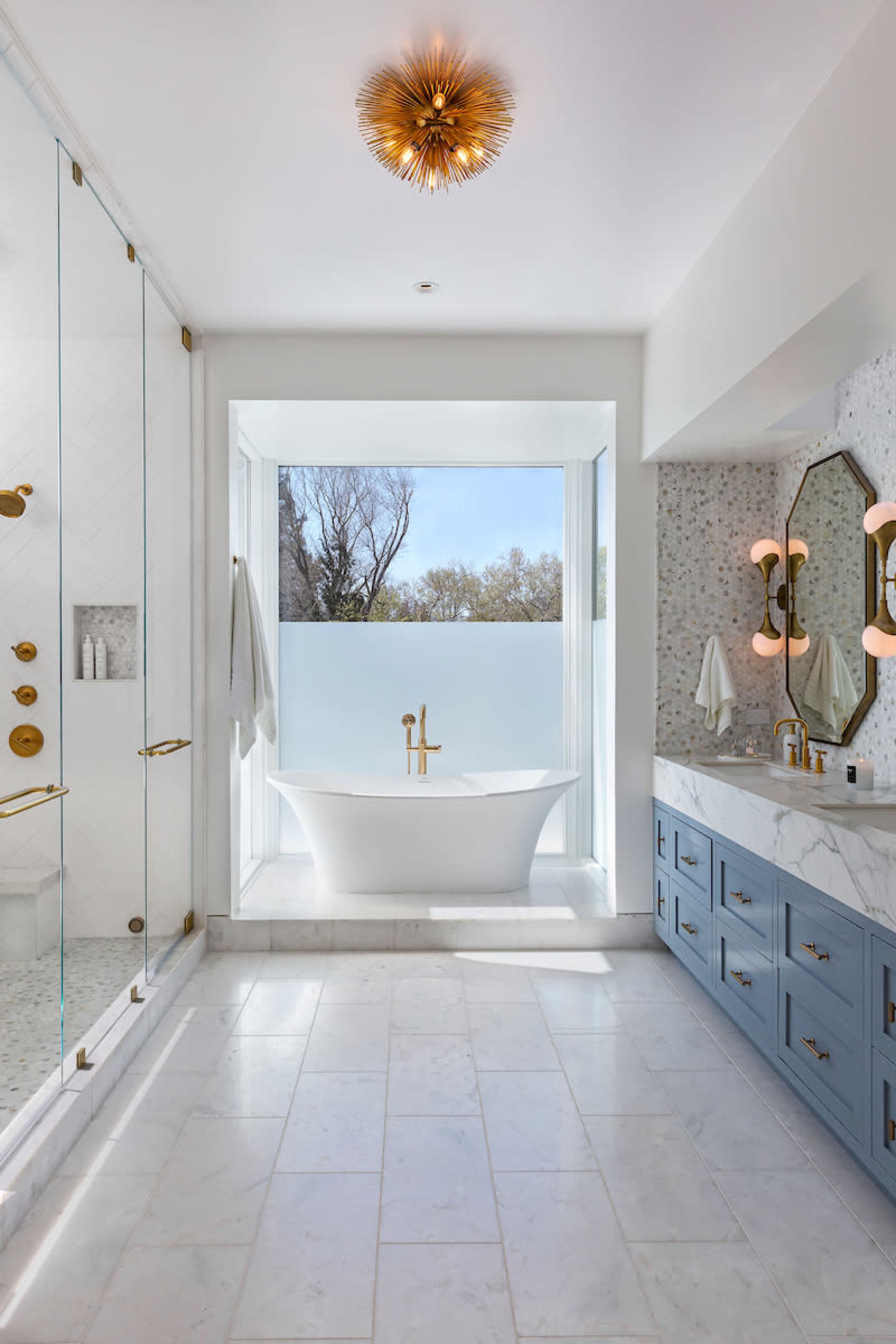 The bathroom features a freestanding bathtub next to a large window, a glass shower enclosure, and a dual-sink vanity with blue cabinets.