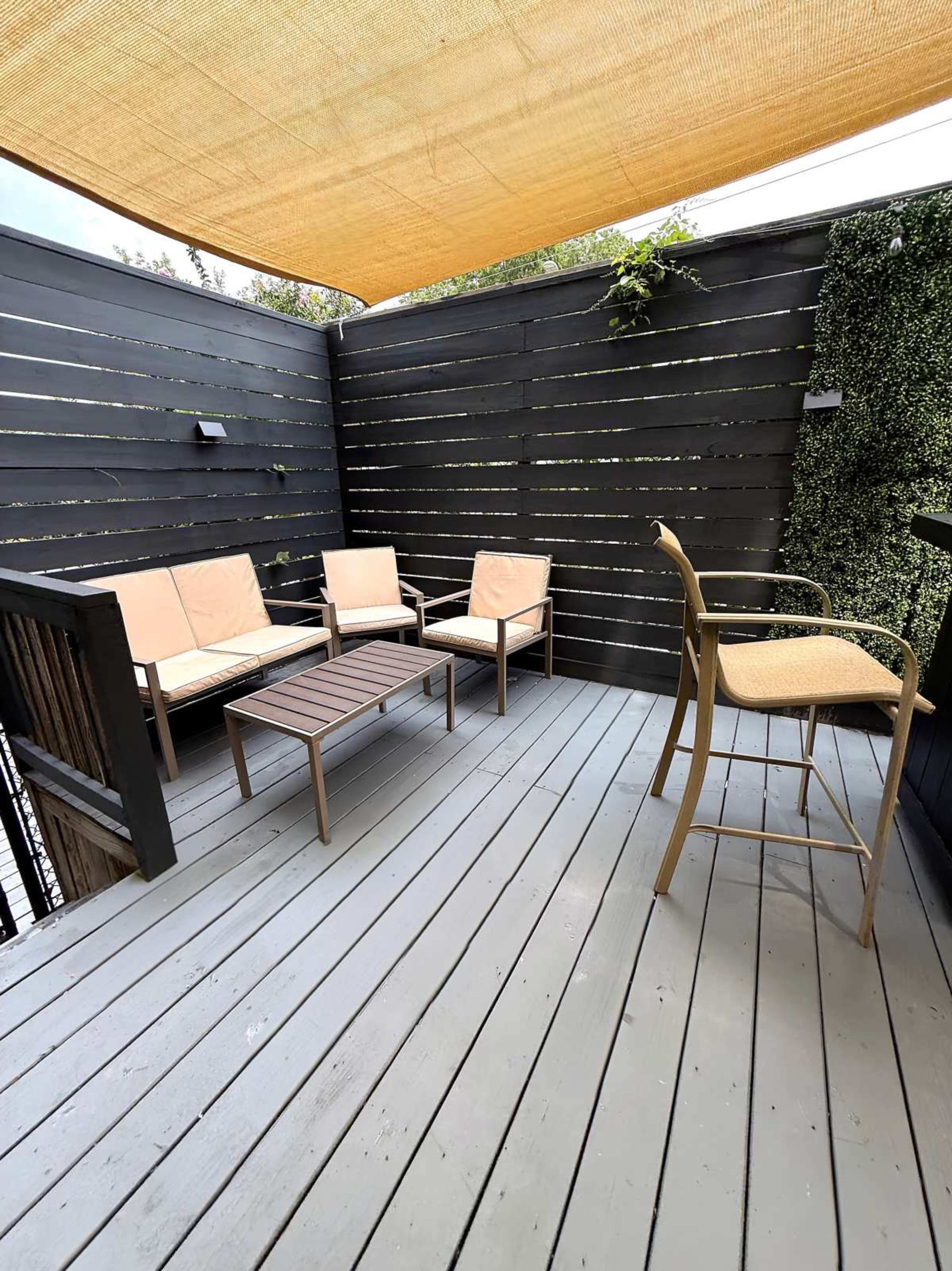 The image shows a small outdoor deck area with seating that includes two armchairs, a coffee table, and a tall bar stool, all under a shaded canopy.