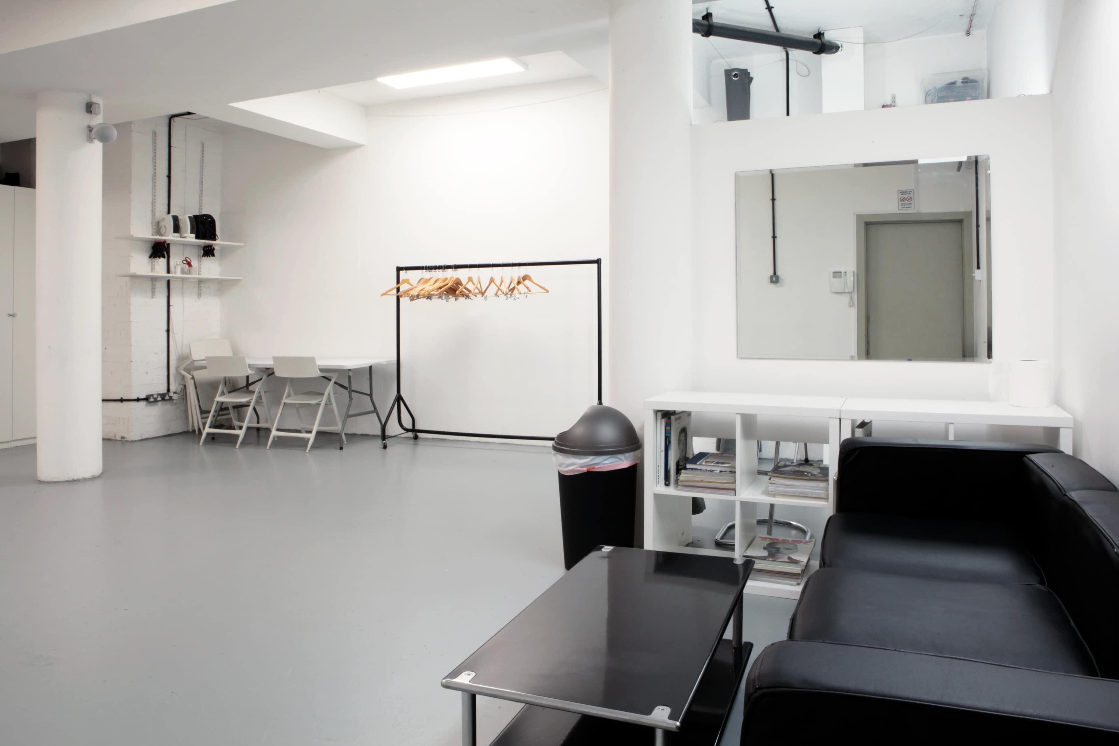 A minimalistic room with white walls, gray flooring, a black couch, a glass table, a clothes rack with hangers, and a mirror on the wall.