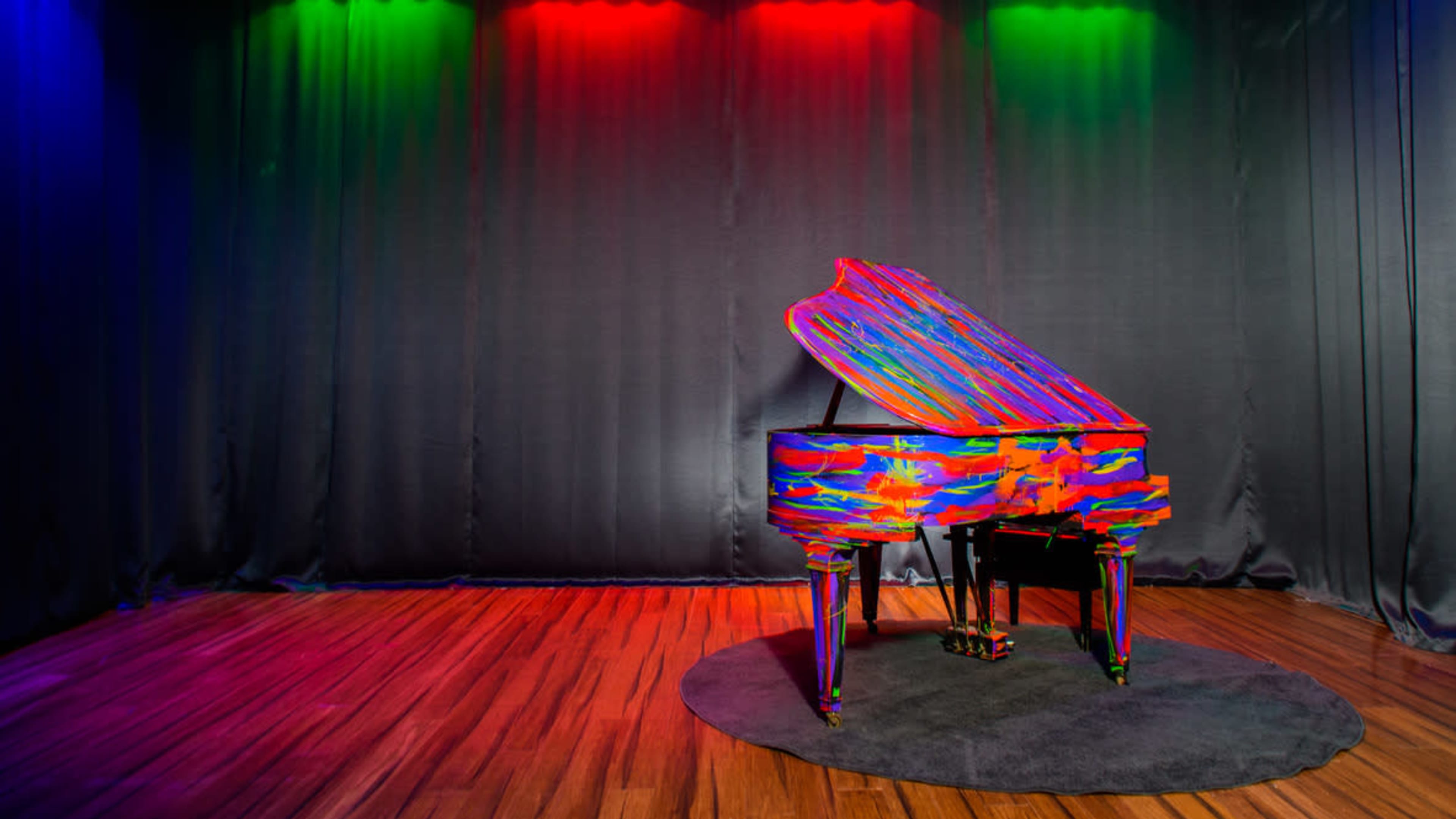A brightly painted grand piano is positioned on a circular rug in front of a dark curtain lit by colored spotlights.