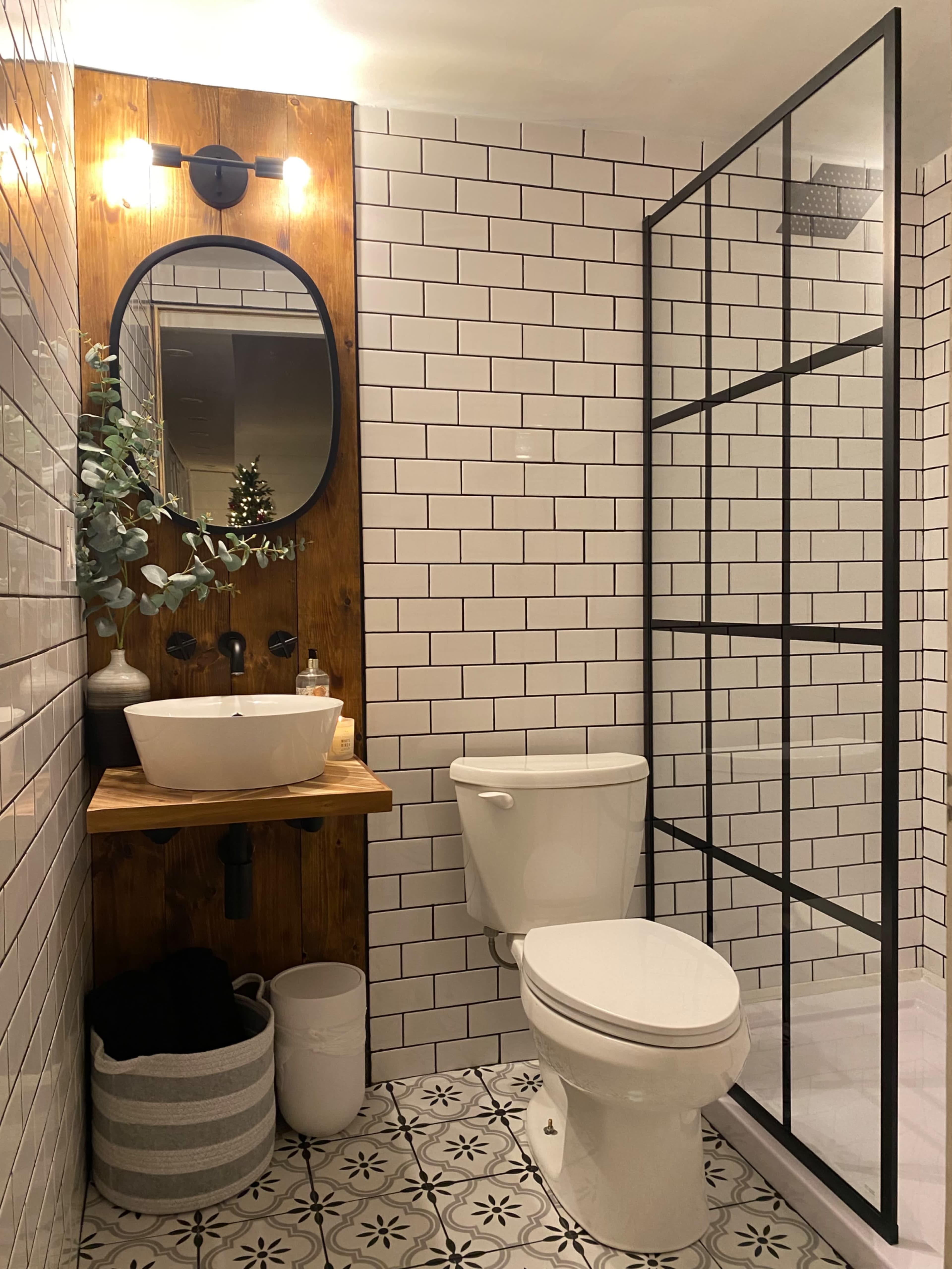 A modern bathroom featuring a wall-mounted sink with a wooden shelf, a black-framed glass shower, white subway tile, and patterned floor tiles.