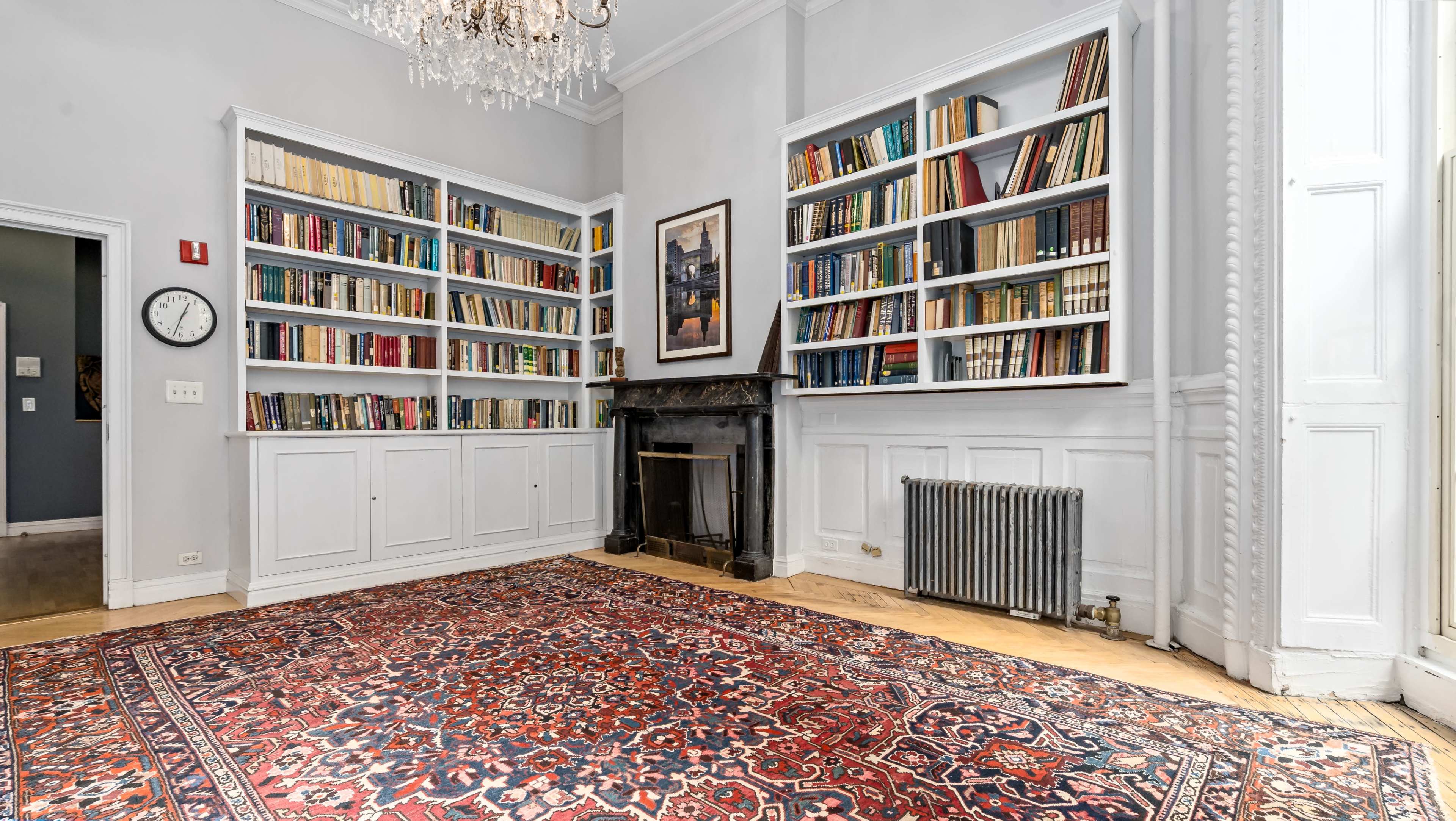 The image shows a room with bookshelves filled with books, a fireplace with a dark mantle, a decorative rug on the floor, and a wall clock.