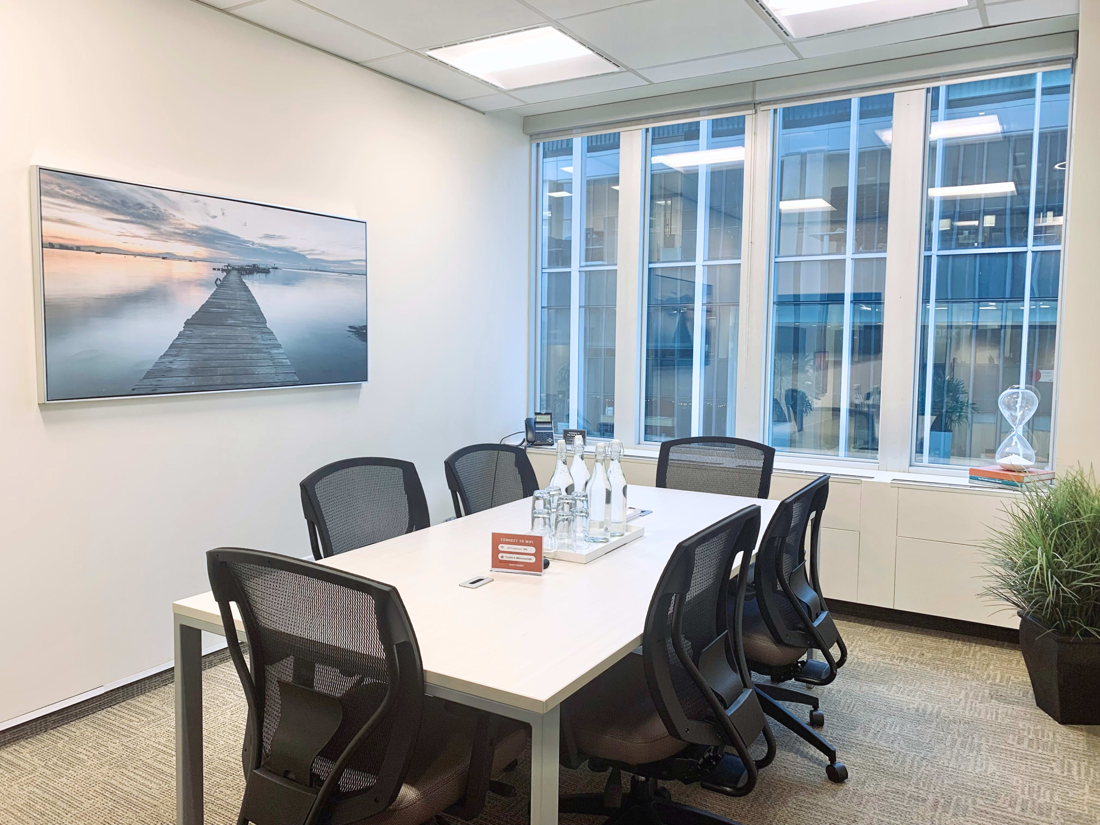 A conference room features a large white table surrounded by ergonomic chairs, with a scenic photograph on the wall and large windows allowing natural light.