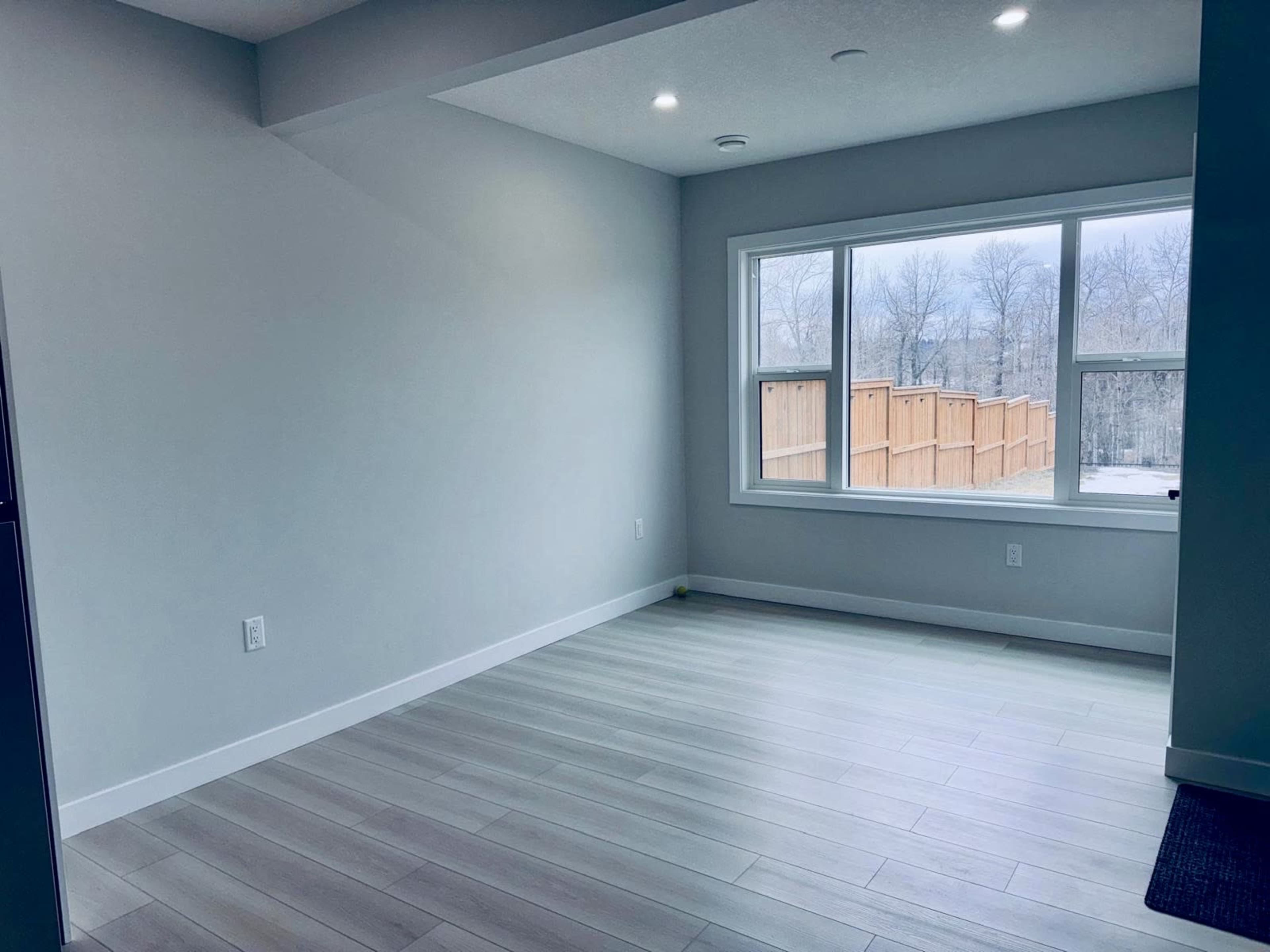 The image shows a vacant room with light-colored walls, wood flooring, and a large window overlooking a fenced outdoor area.
