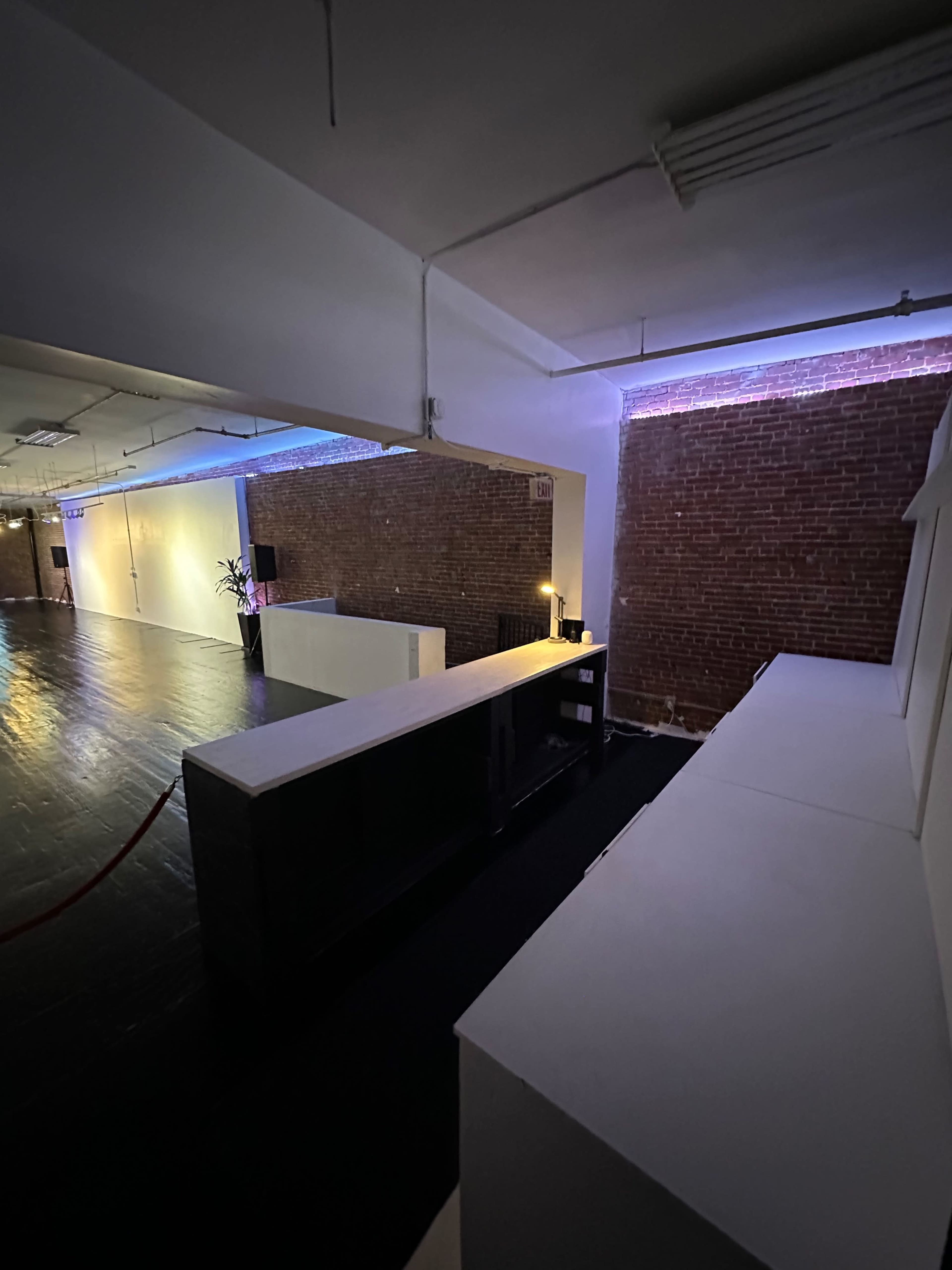 The image shows a spacious room with exposed brick walls, a dark wooden floor, and a white counter along one side, illuminated by soft lighting from above.