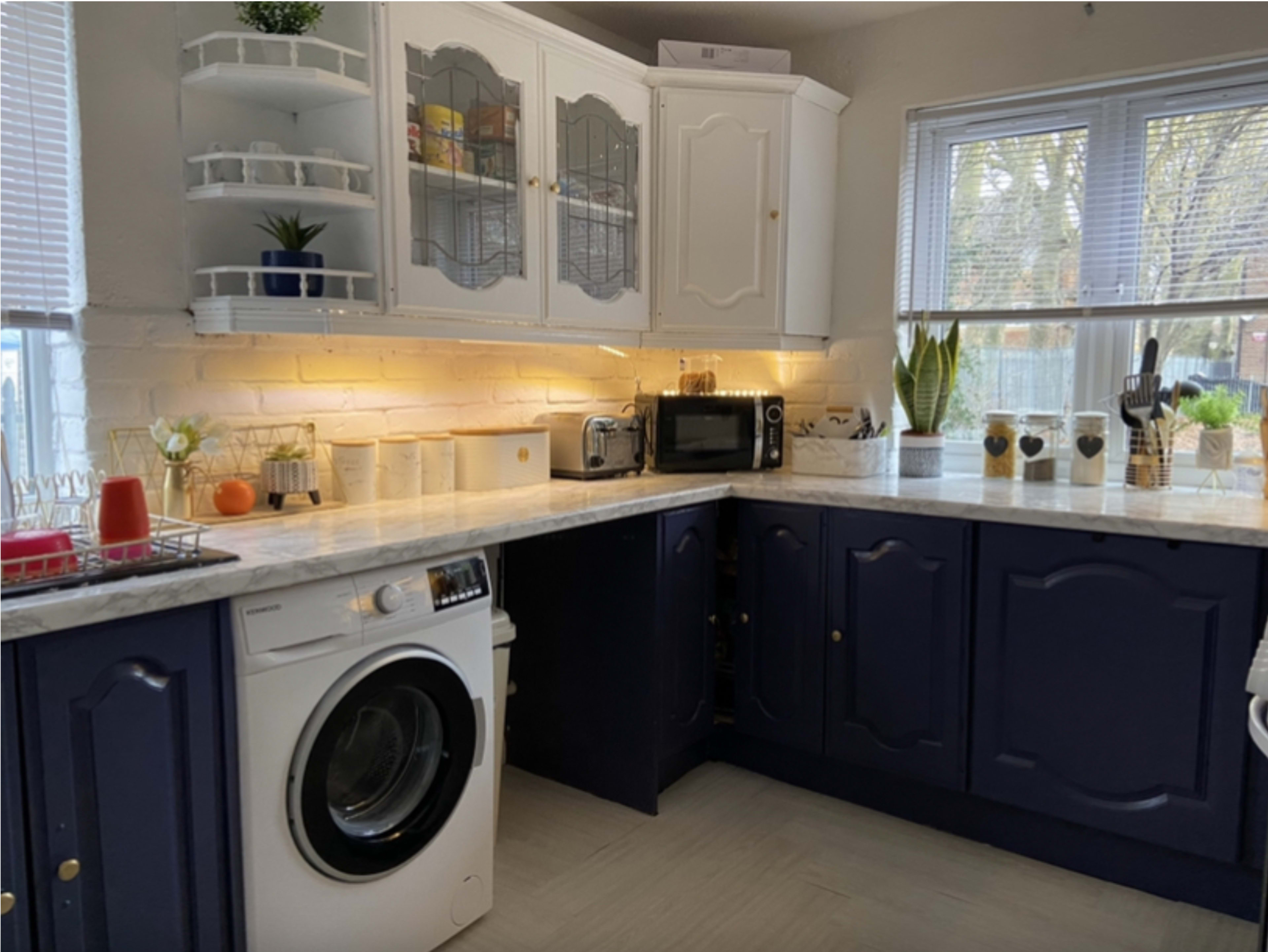 A kitchen with navy blue cabinets, a marble countertop, a washing machine, and various appliances including a microwave and a coffee maker.