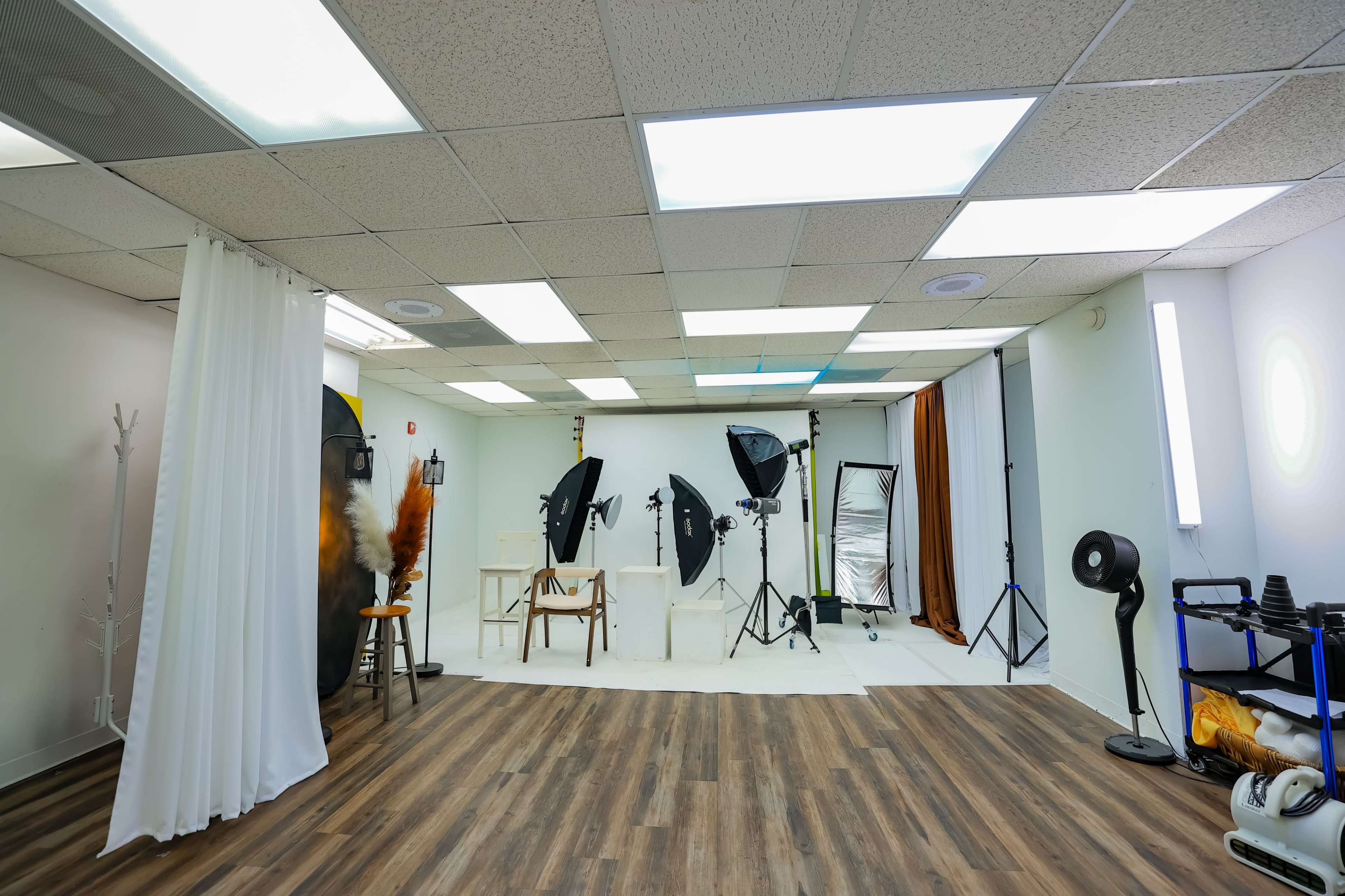 The image shows a photography studio equipped with various lighting setups, backdrops, and props arranged in a spacious room with wooden flooring.