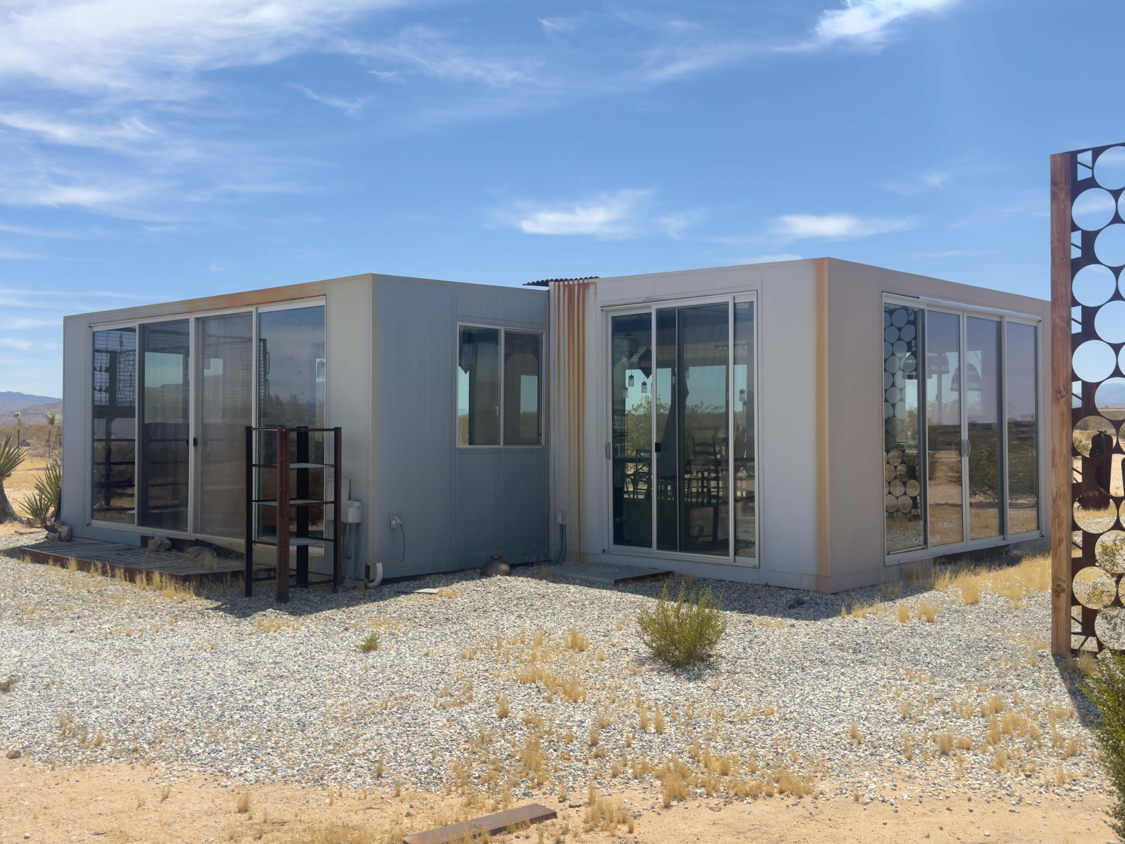 The image shows a modern, angular structure with large glass windows and a metallic exterior, situated on a gravelly terrain in a desert landscape.