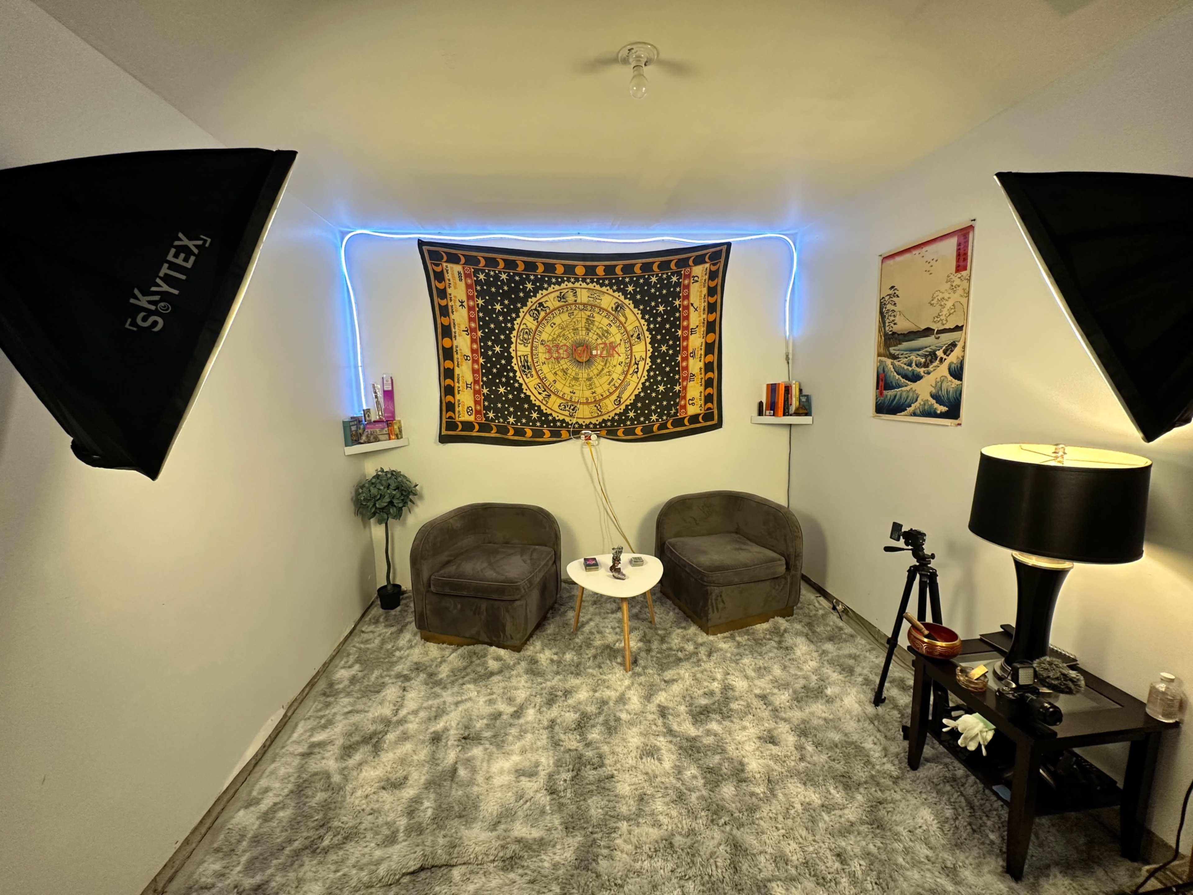 The image shows a small, well-lit room with two gray chairs, a small white table, a decorative tapestry hanging on the wall, and a lamp beside a low table.