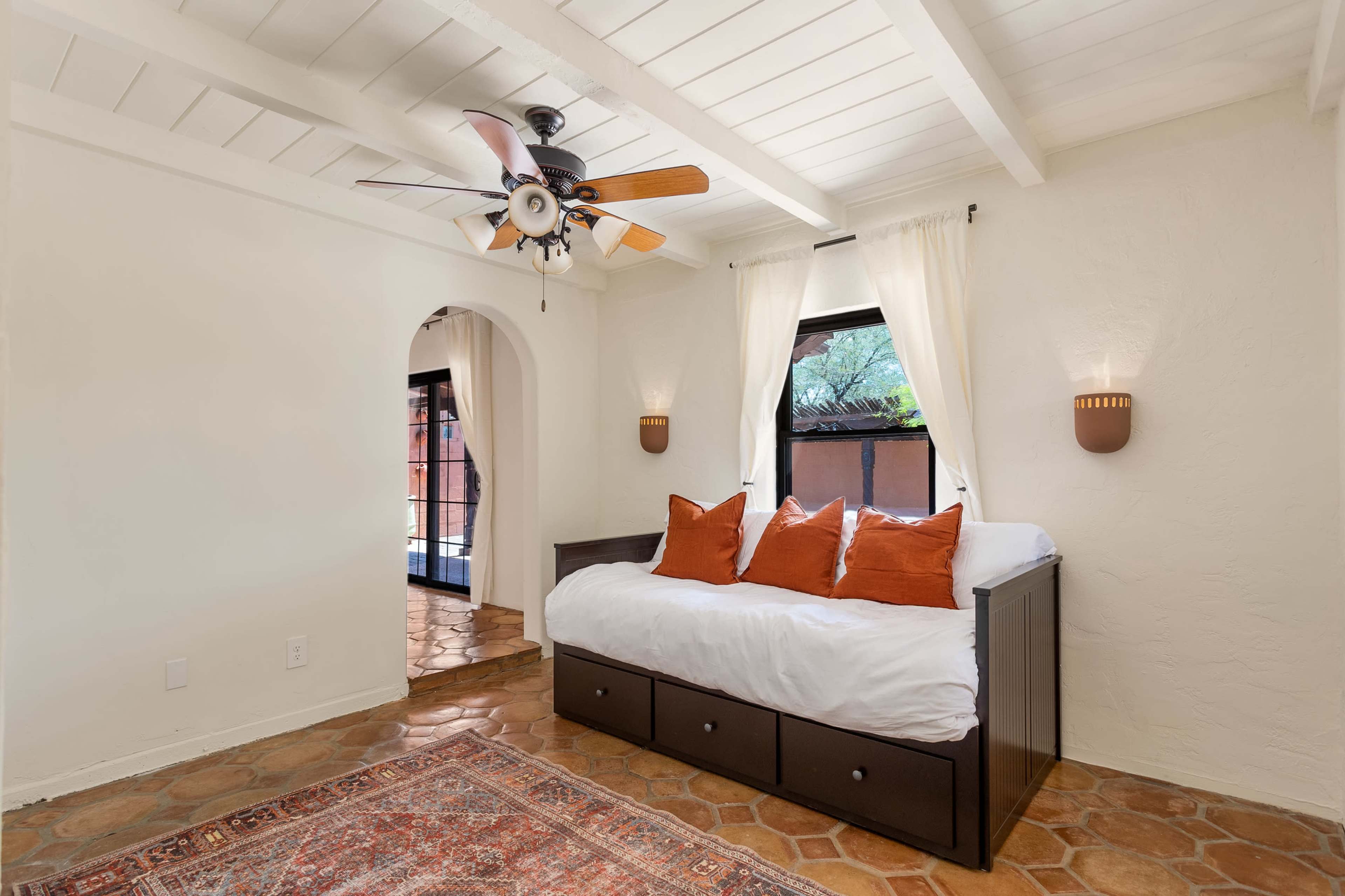 The room features a daybed with orange pillows, a ceiling fan, and a terracotta tiled floor, illuminated by natural light through two windows.