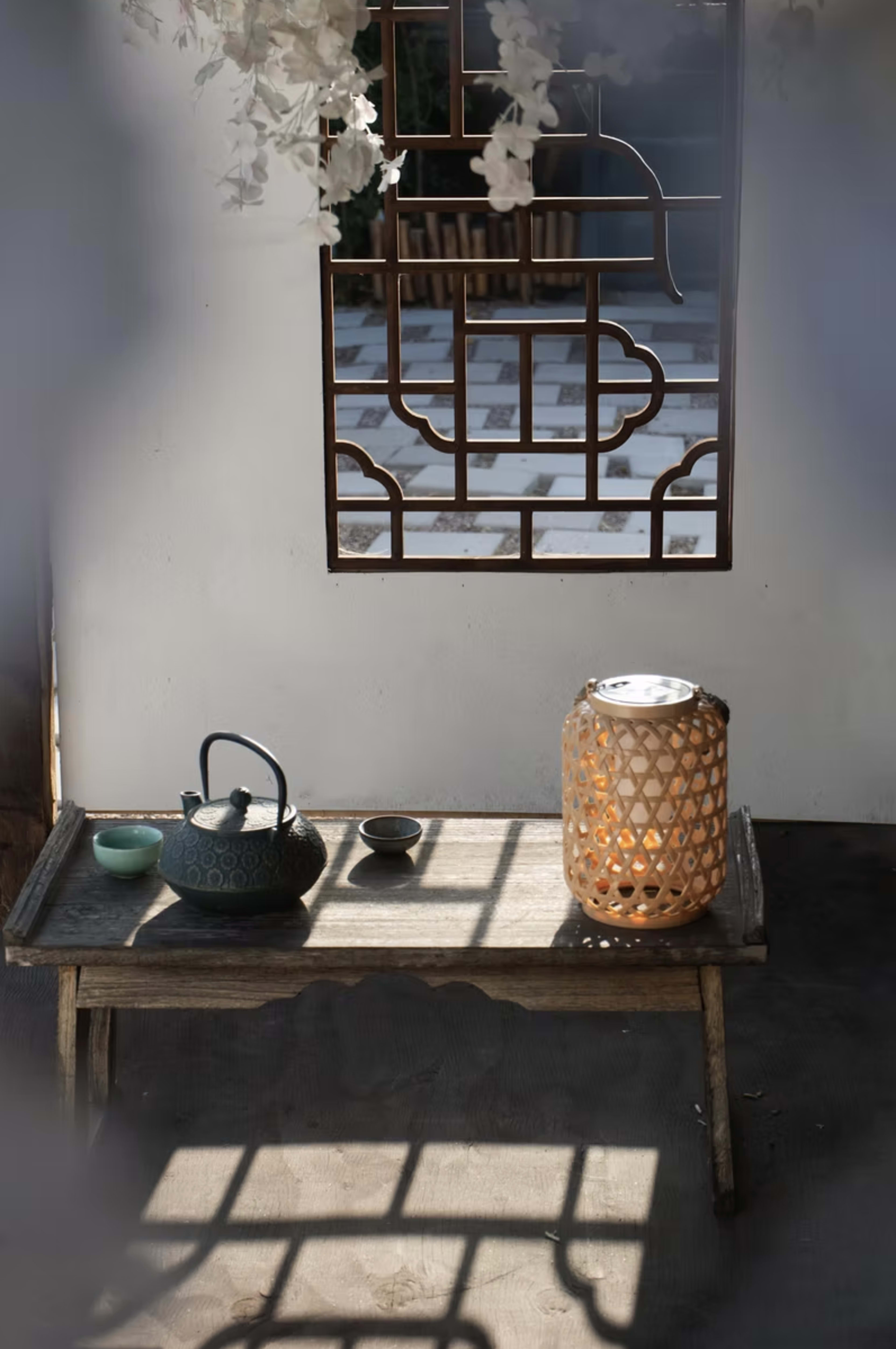 A wooden table with a teapot, a small bowl, and a lantern sits in front of a decorative screen, illuminated by soft light.
