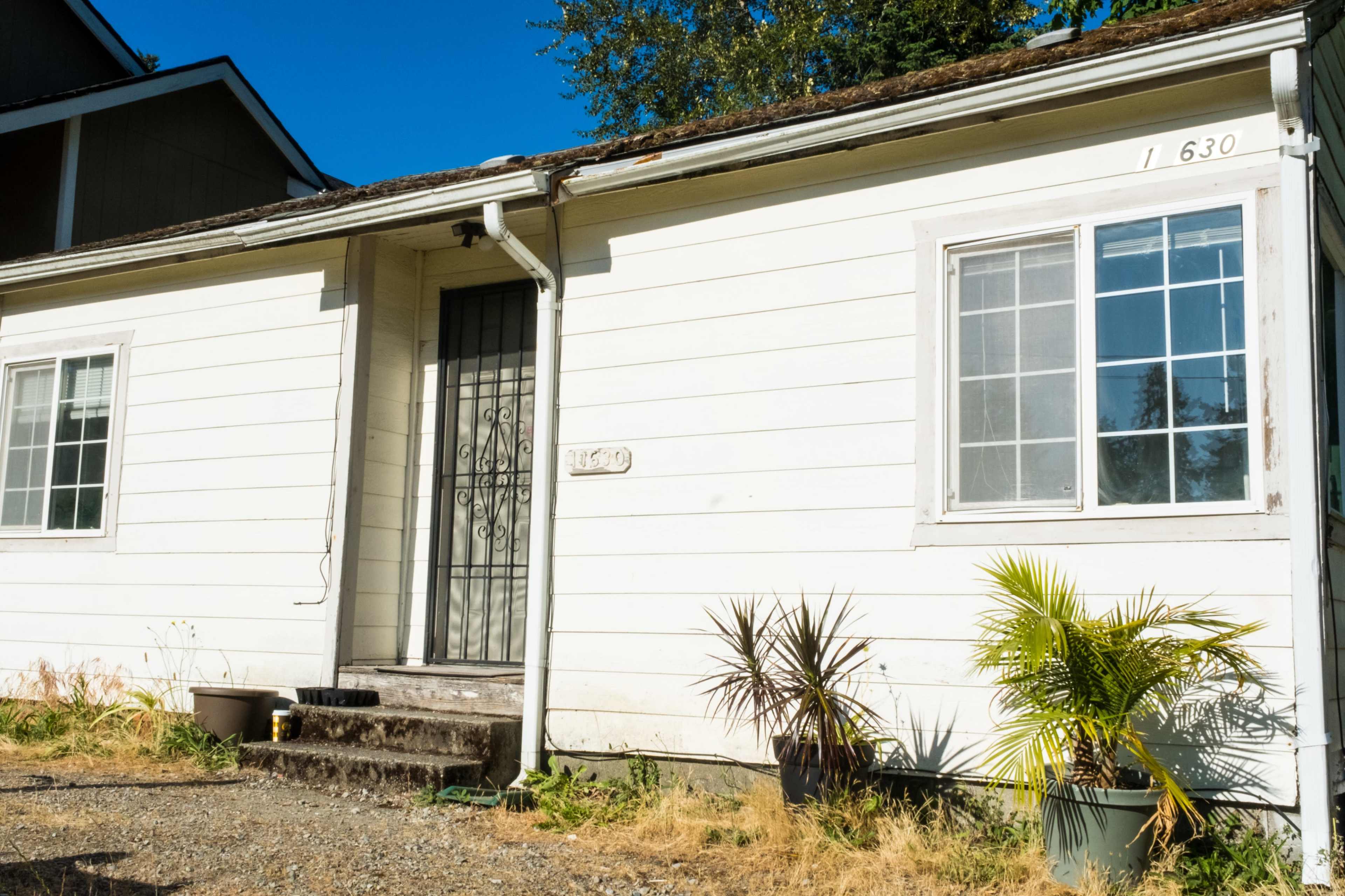 A small, white single-story house with a front porch, a metal security door, and two windows on one side, surrounded by potted plants and dry grass.