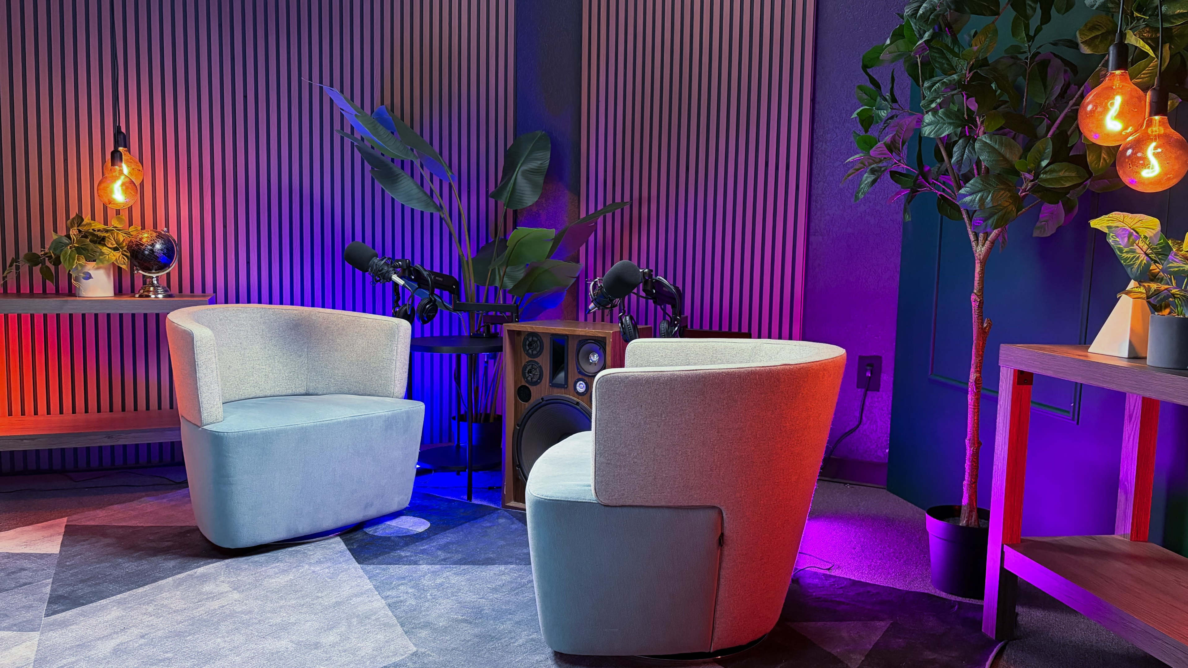 The image depicts a cozy recording studio setup with two chairs, microphones on stands, and decorative plants against a backdrop of patterned walls and colorful lighting.