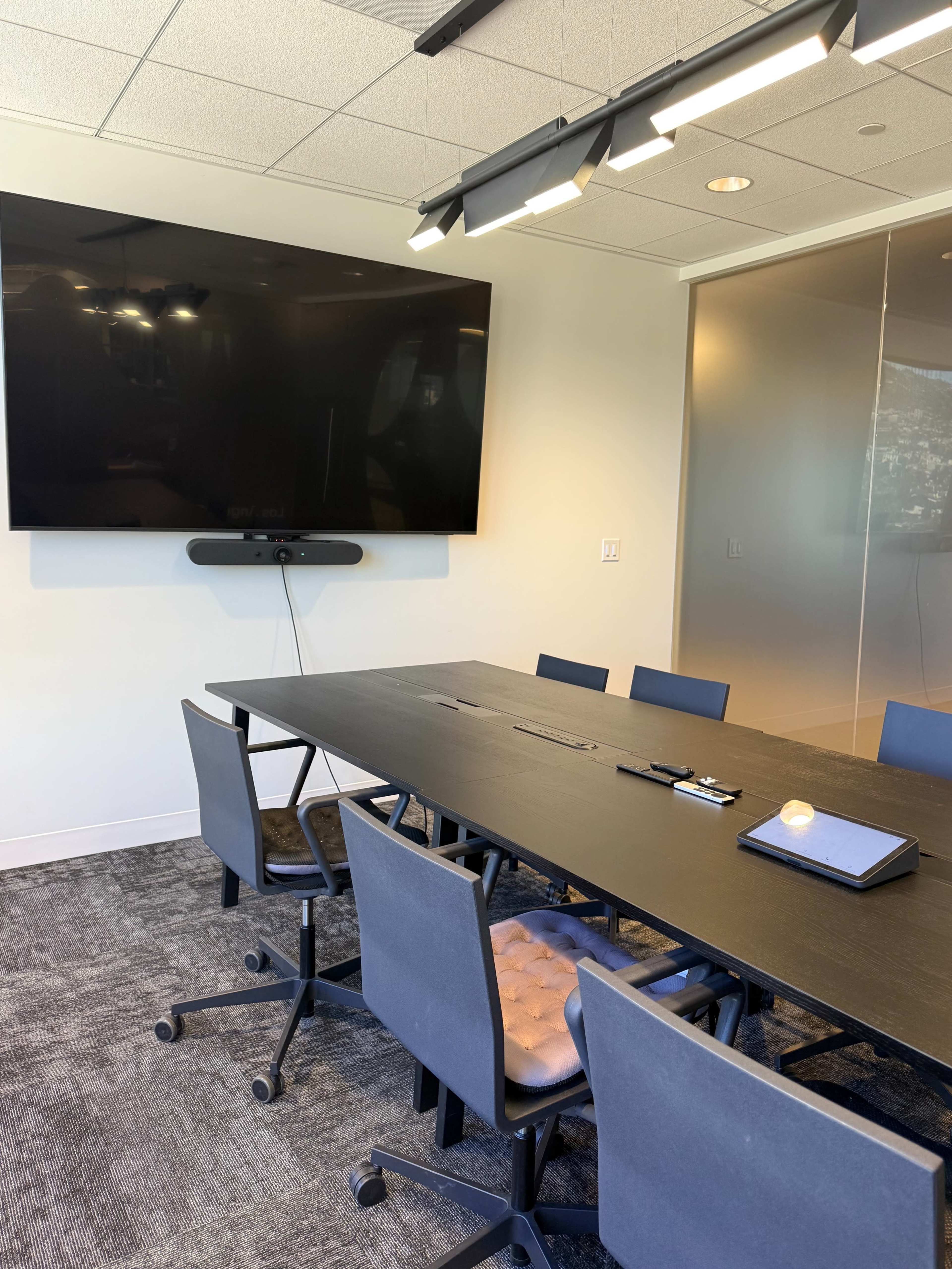 A conference room features a large black table surrounded by several chairs, with a wall-mounted television and a window in the background.