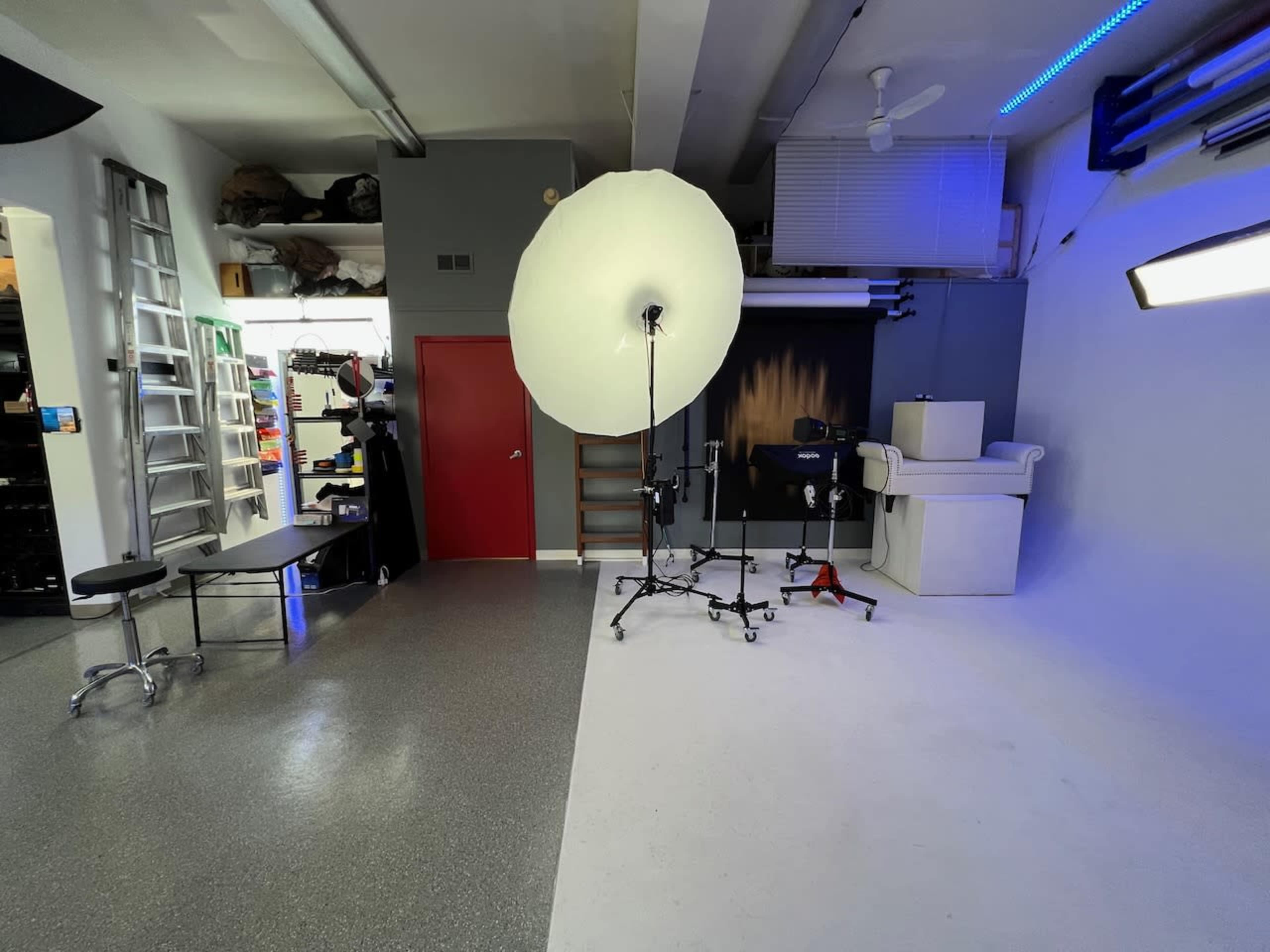 A photography studio with a large light umbrella on a stand, a white backdrop, and various equipment placed in the corners.