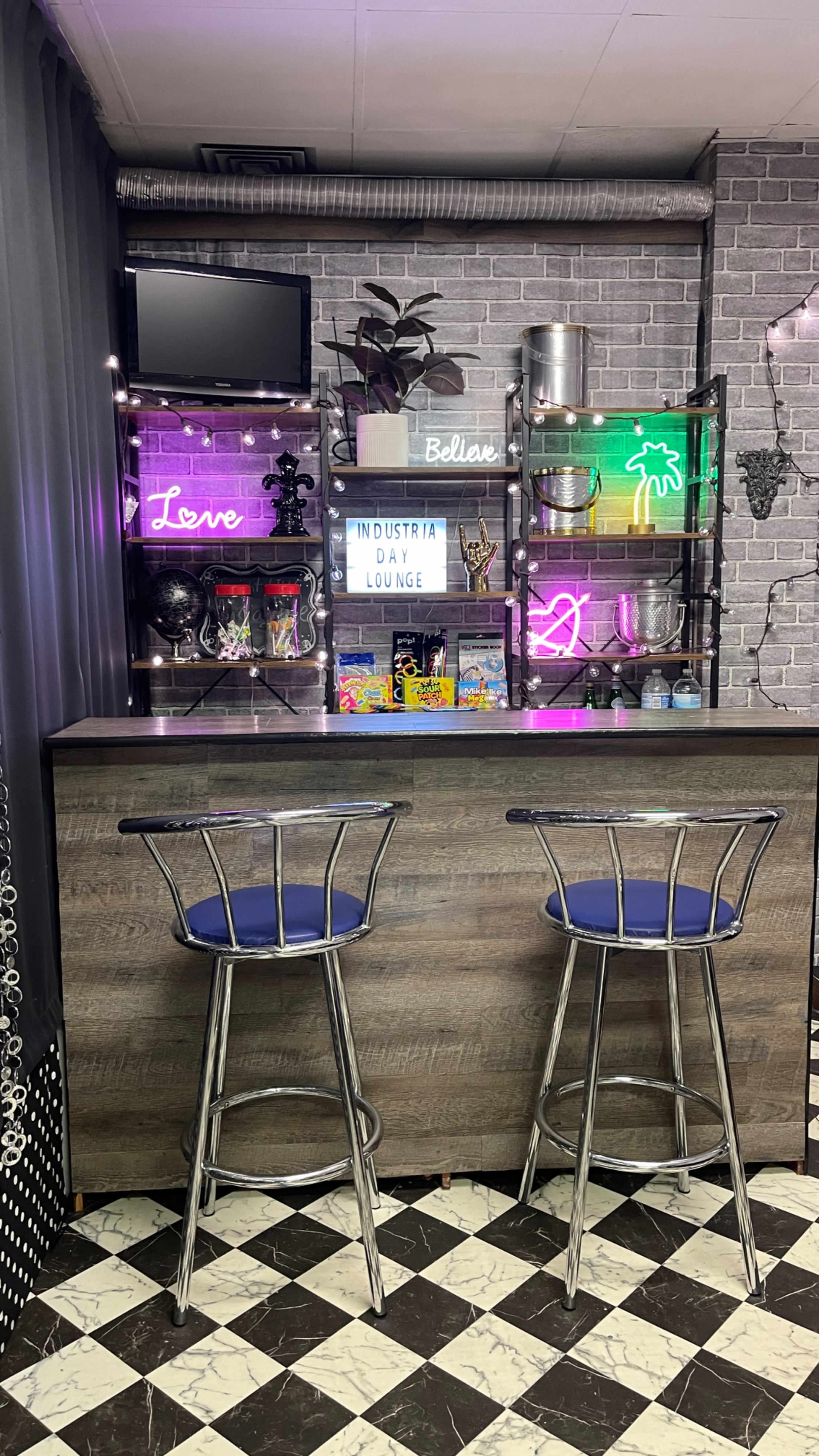 The image shows a modern bar area featuring a countertop with two stools, colorful neon signs, shelves filled with decor and drinks, and a checkered floor.