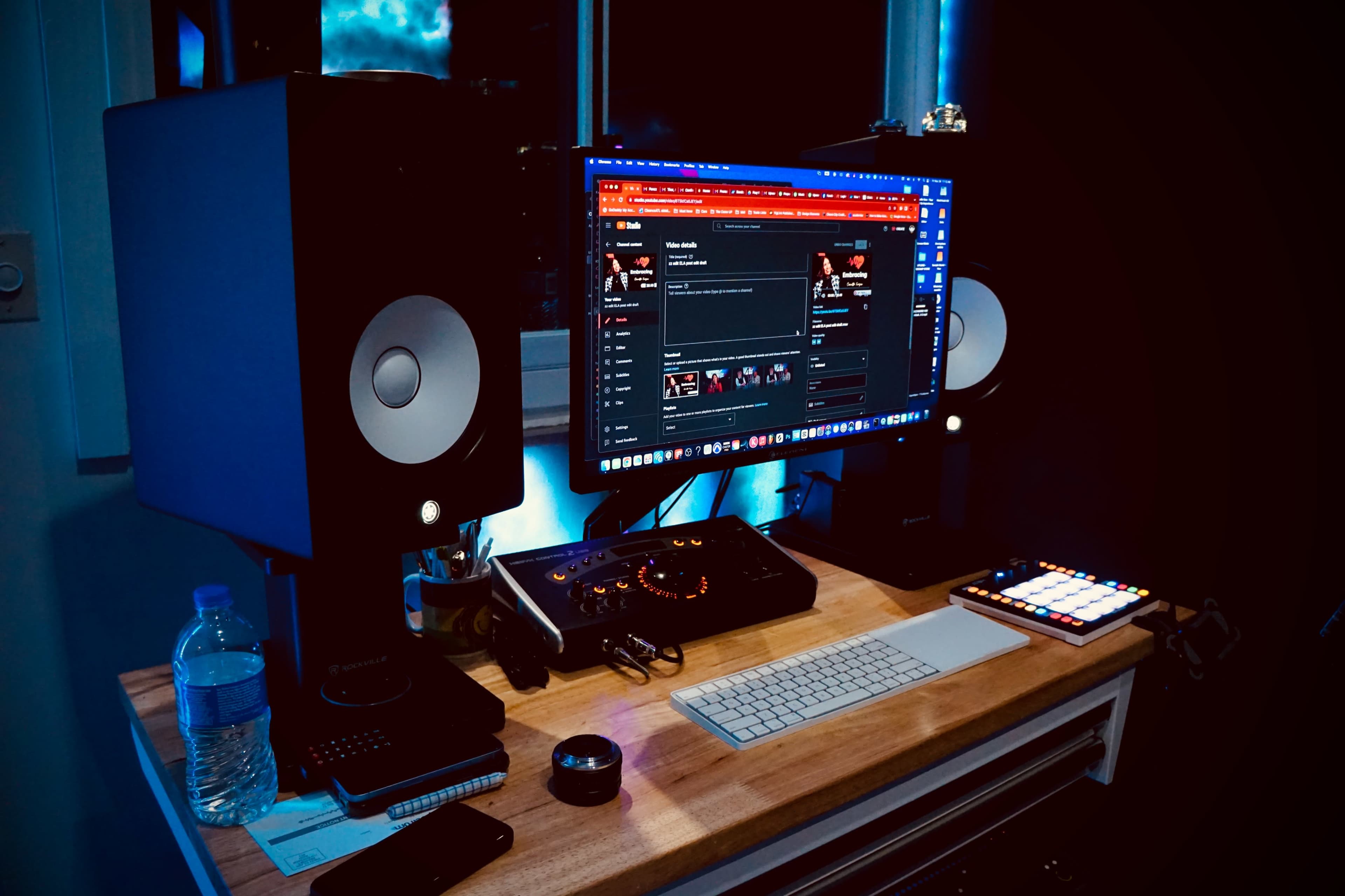 A music production setup is shown with a computer monitor, audio speakers, a DJ controller, and various devices arranged on a wooden desk.