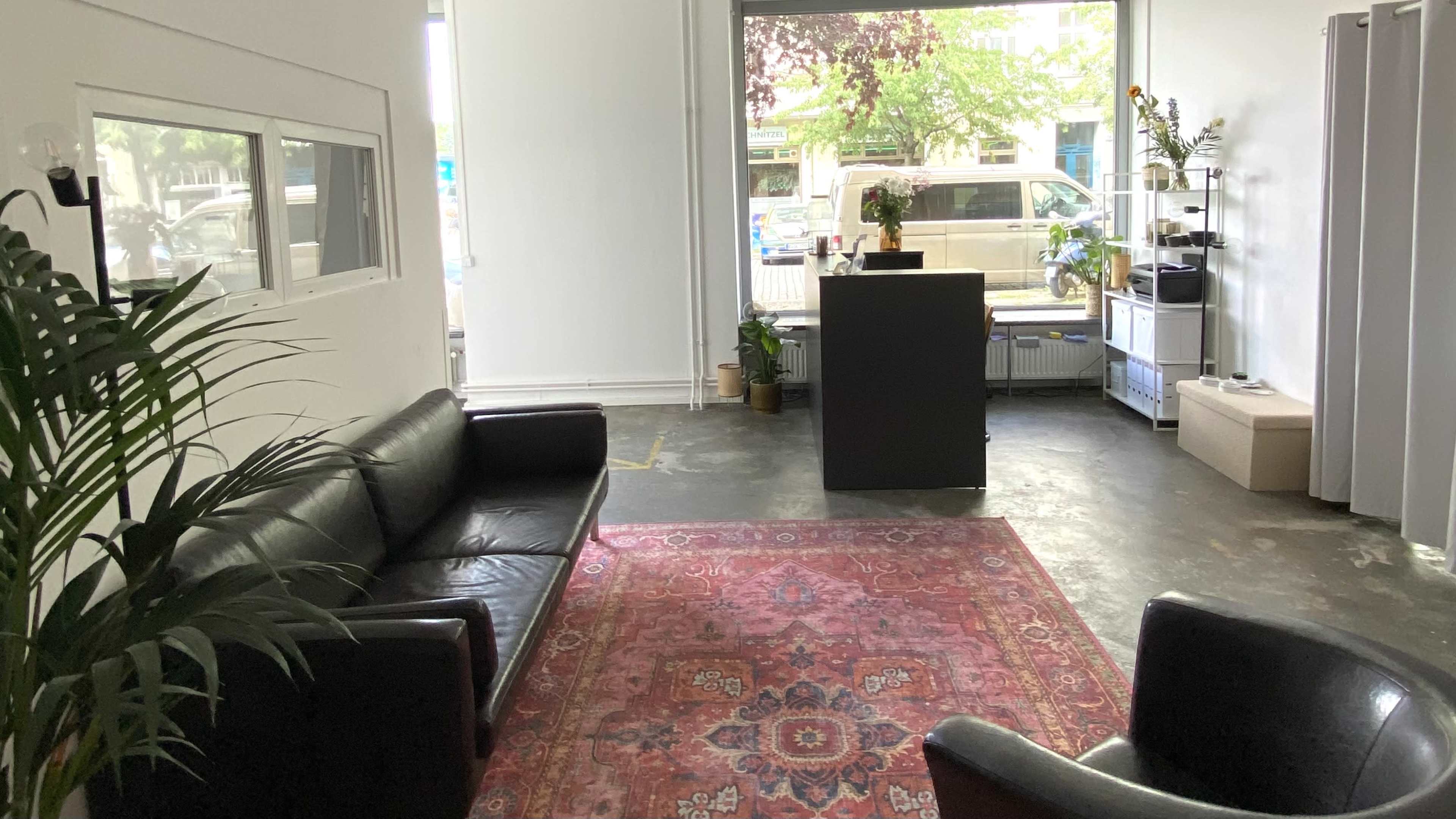 The image shows a spacious interior with a black leather couch and armchair, a rectangular desk, and large windows facing the street, revealing an outdoor view.