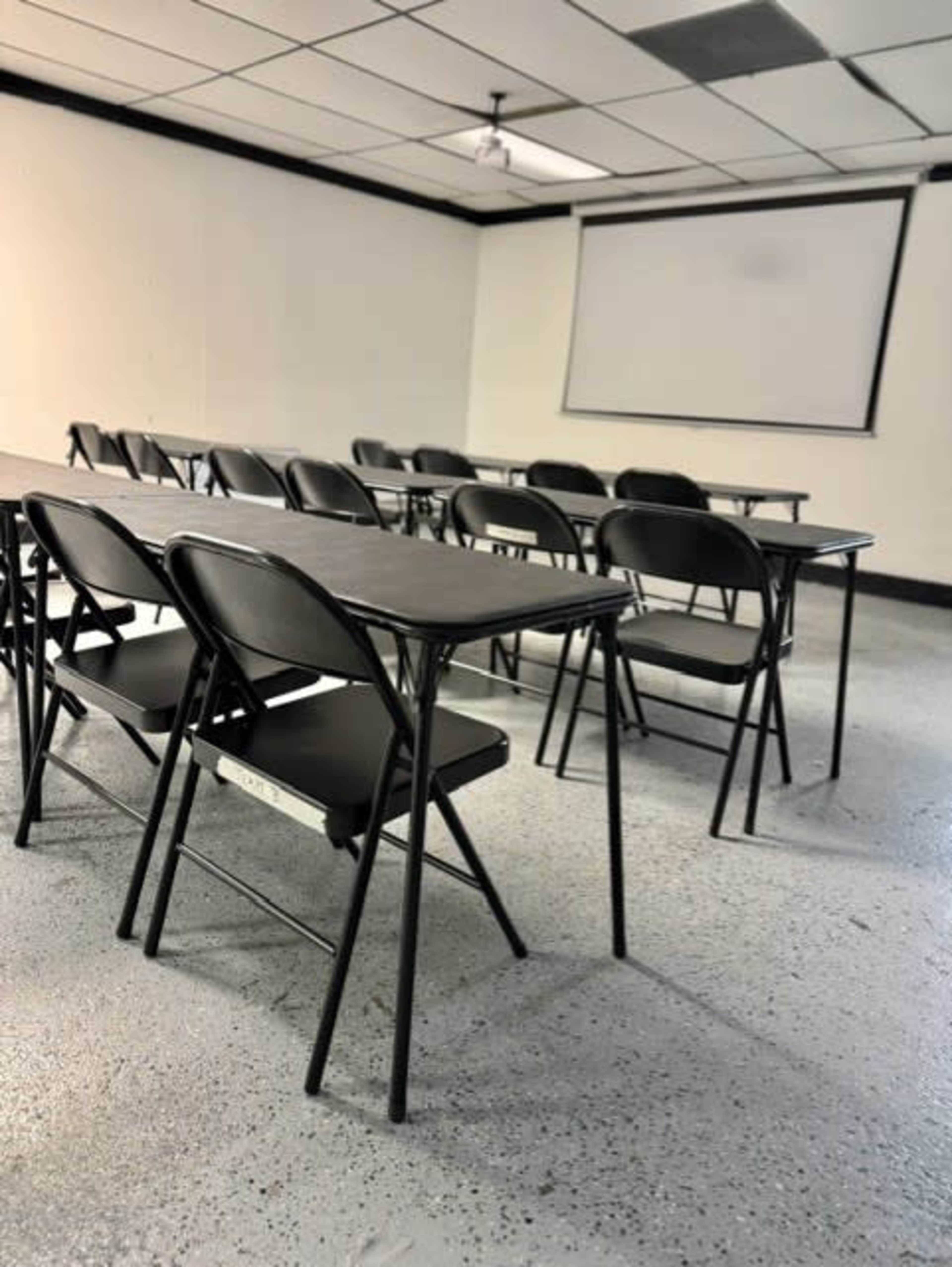 The image shows an empty classroom with several rows of black folding chairs and tables, facing a blank projection screen on the wall.