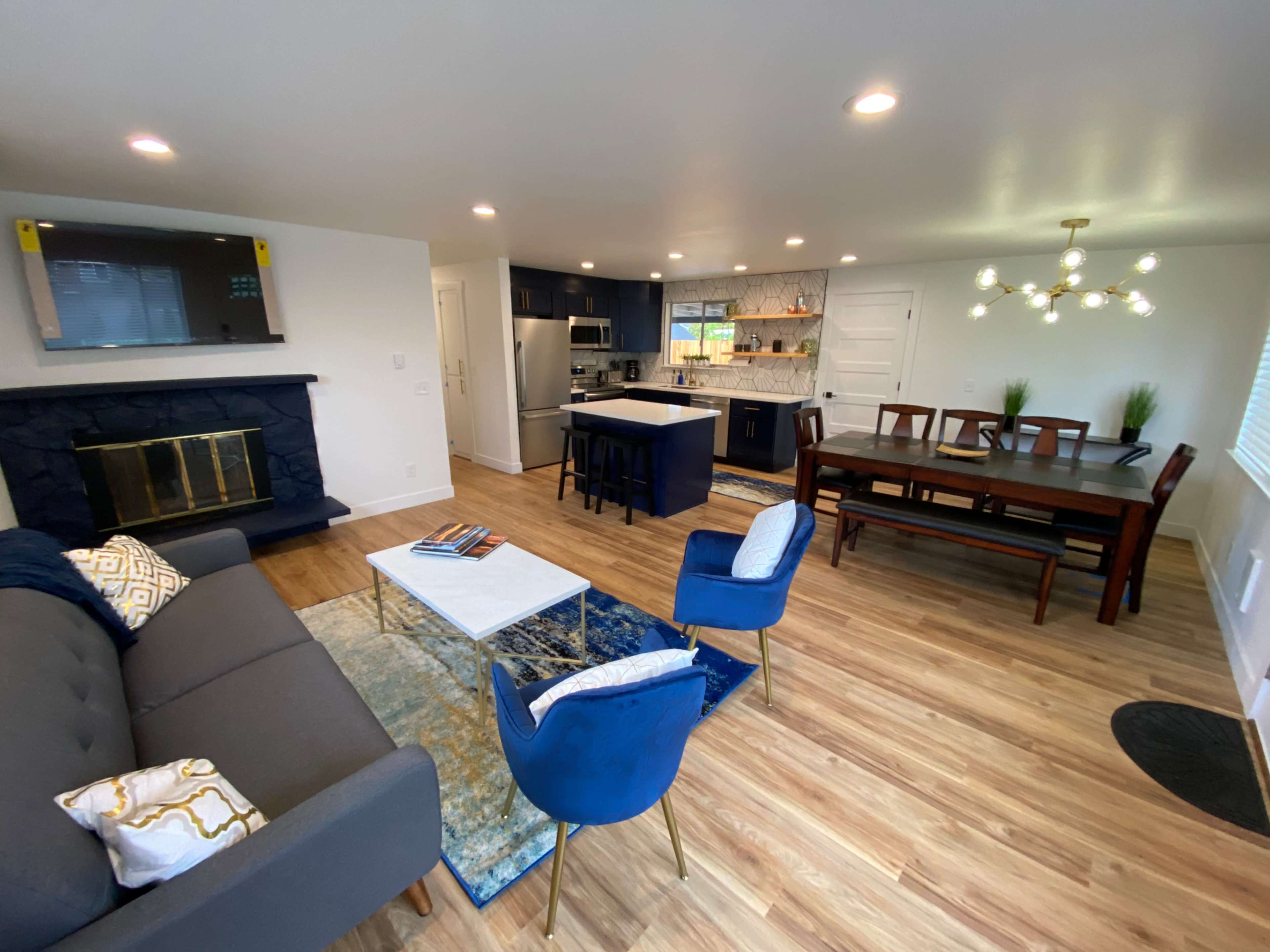 A modern living space featuring a gray sofa, a wooden dining table, a kitchen area with navy blue cabinets, and a decorative fireplace.