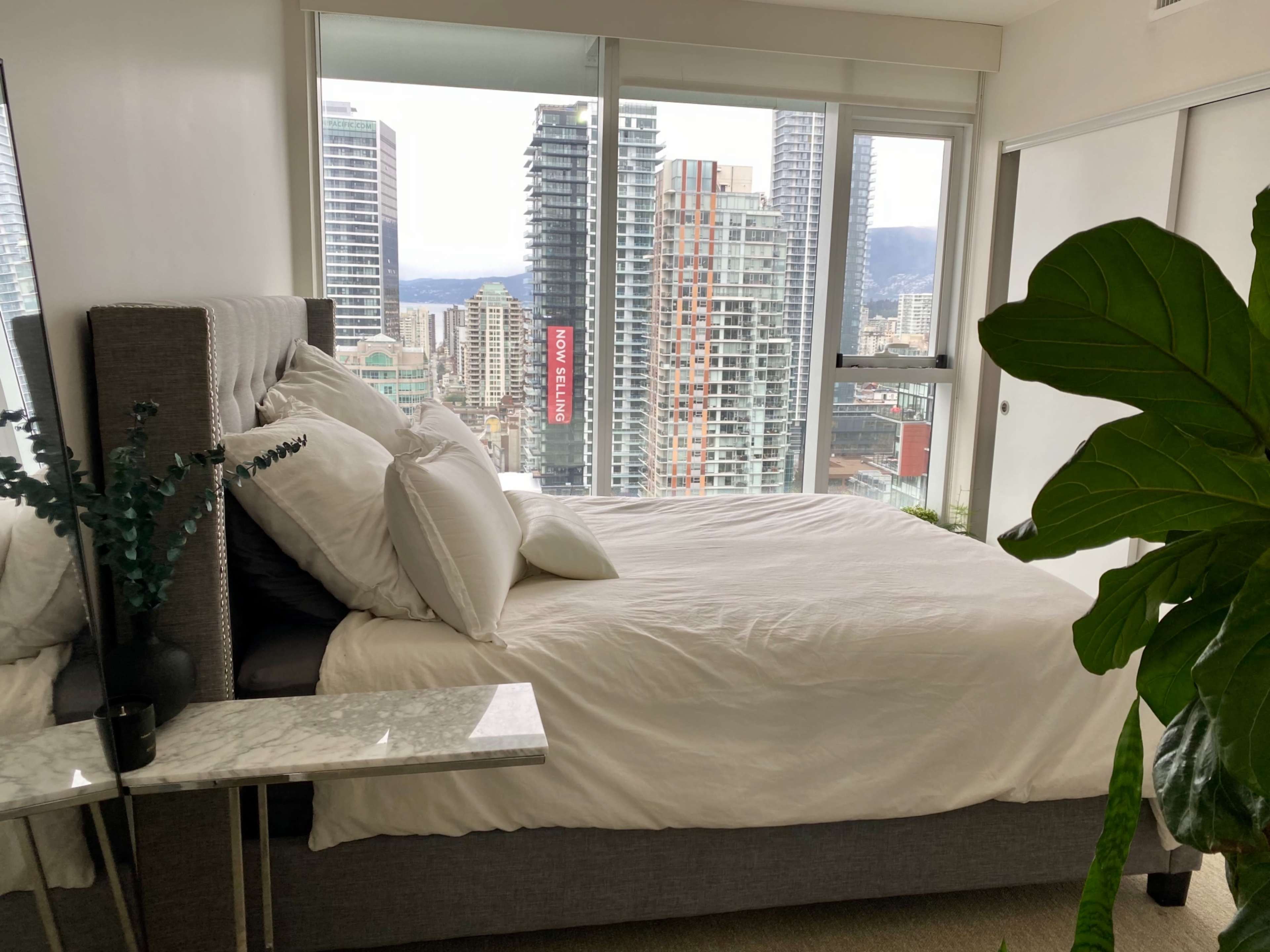 A neatly made bed sits beside a large window overlooking a city skyline with tall buildings.