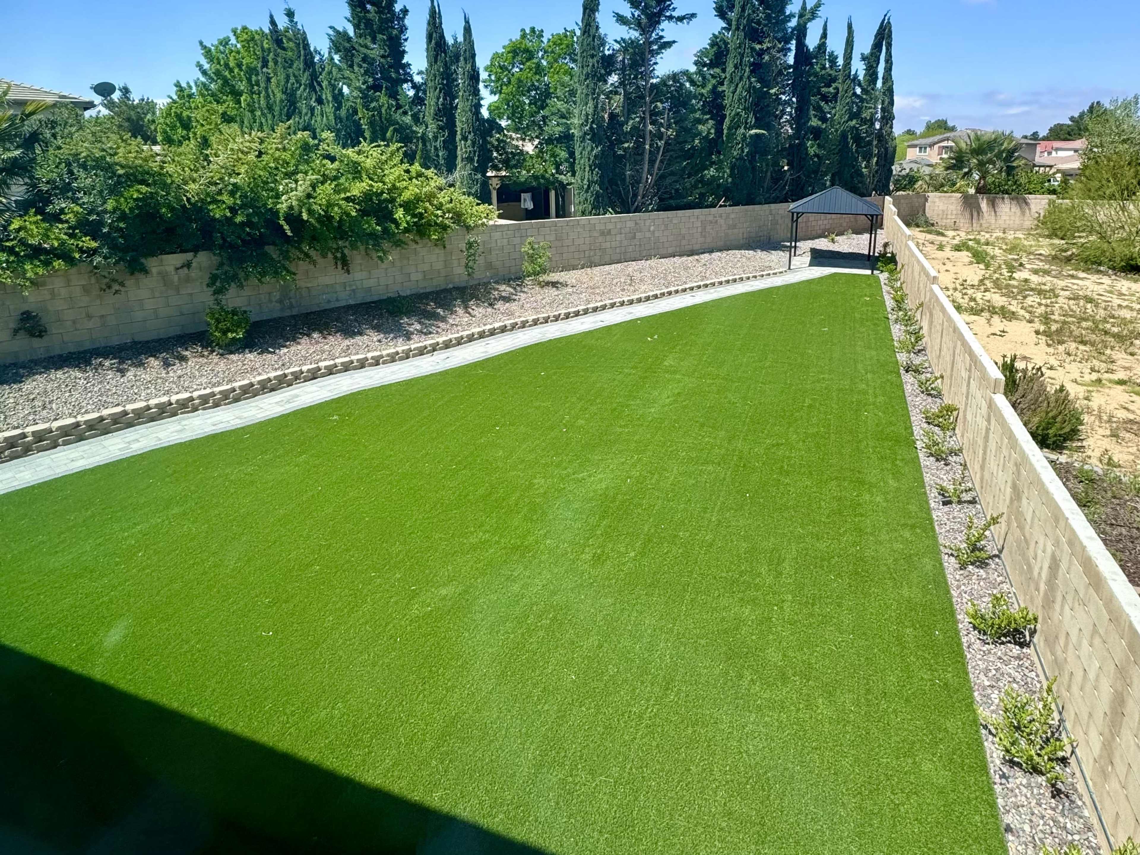 A landscaped outdoor area with artificial turf, bordered by stone pathways and plants, set against a backdrop of trees and a low wall.