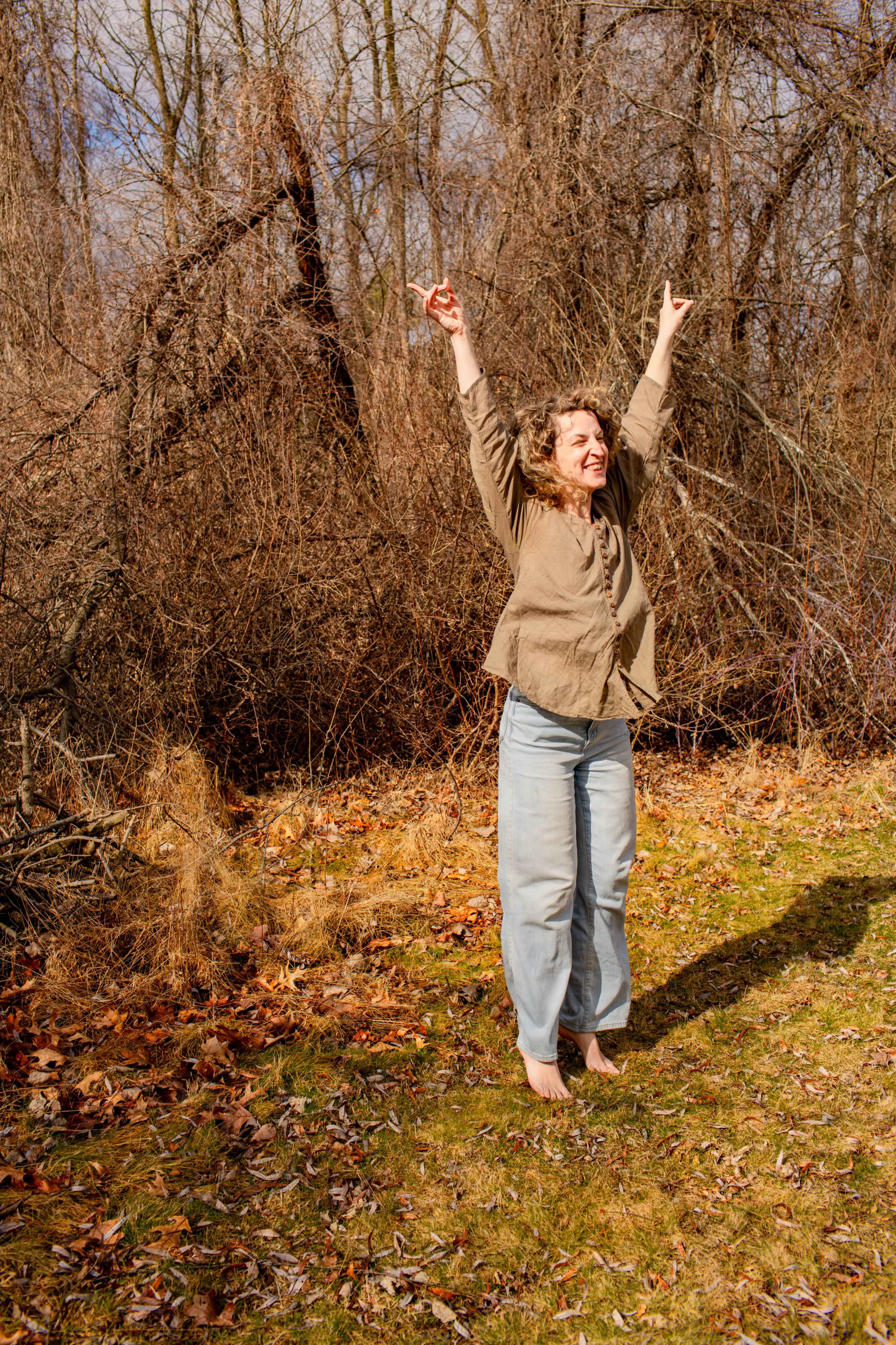 A woman stands with her arms raised in a grassy area surrounded by bare trees and shrubs.