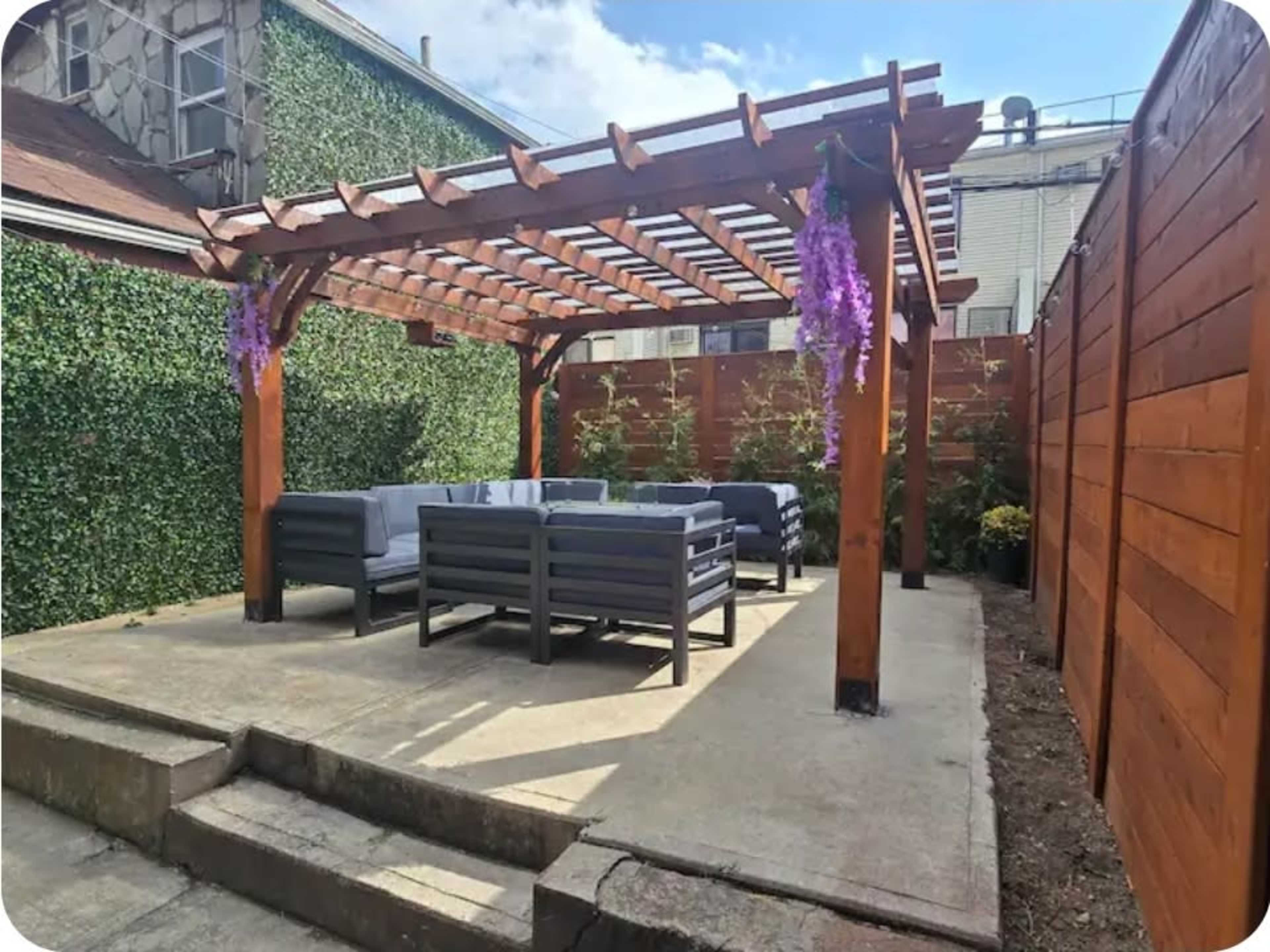 A wooden pergola with purple wisteria flowers covers a seating area featuring gray outdoor furniture on a concrete patio surrounded by wooden fencing and greenery.