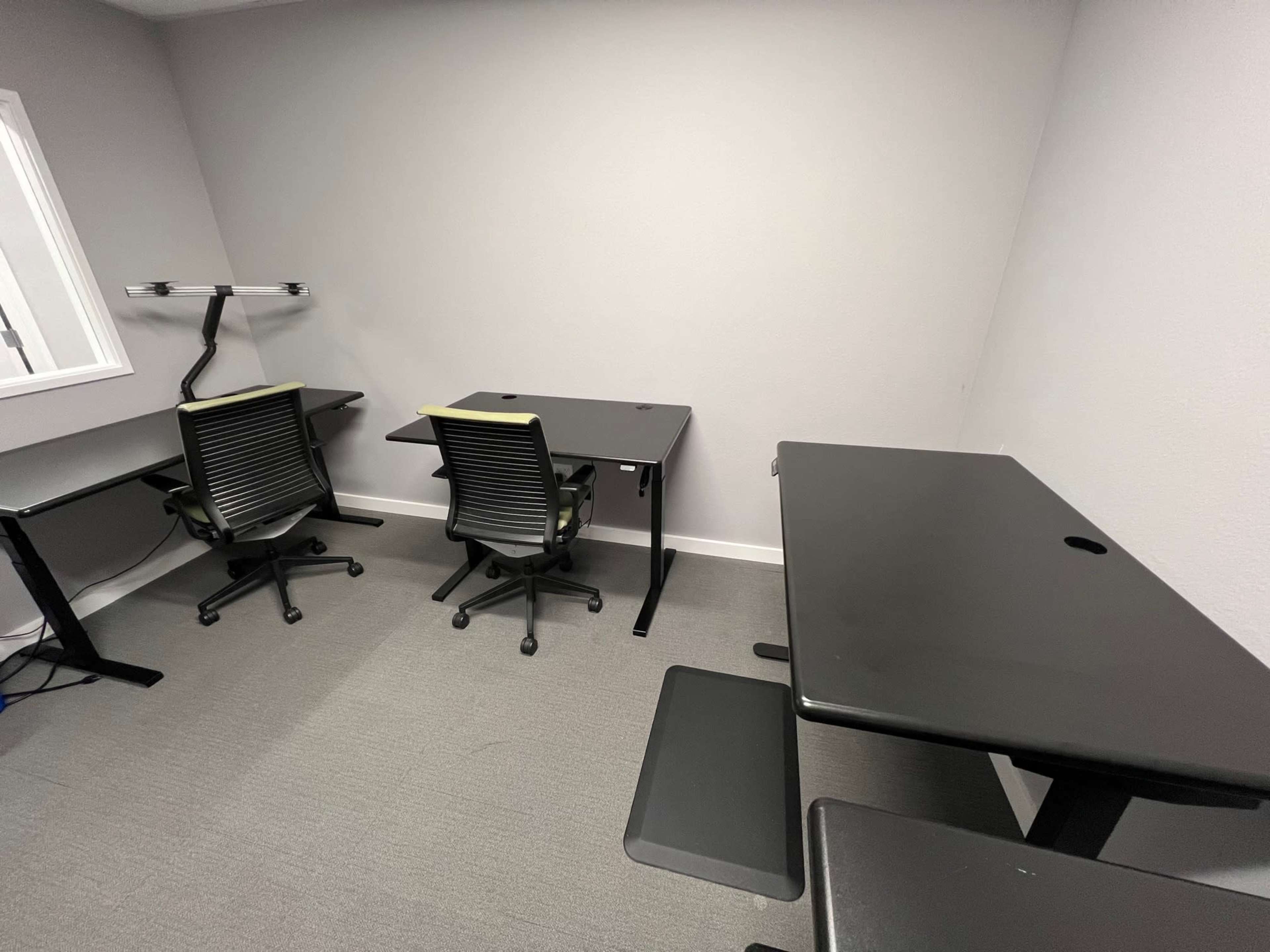 A minimalist office space featuring two black desks and matching ergonomic chairs against a gray wall.