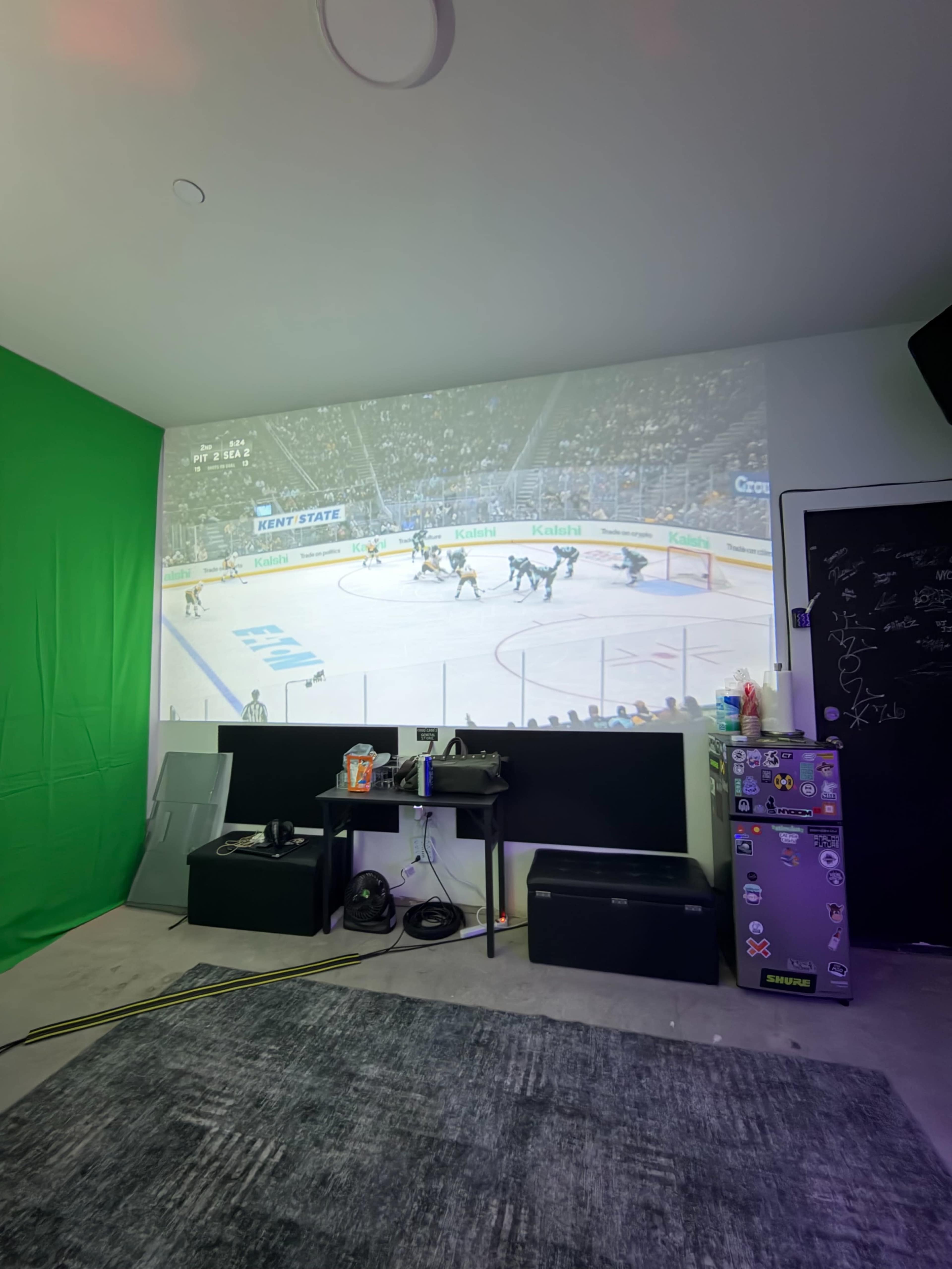 A hockey game is being projected onto a wall in a room with green screen material and various items on a cabinet.