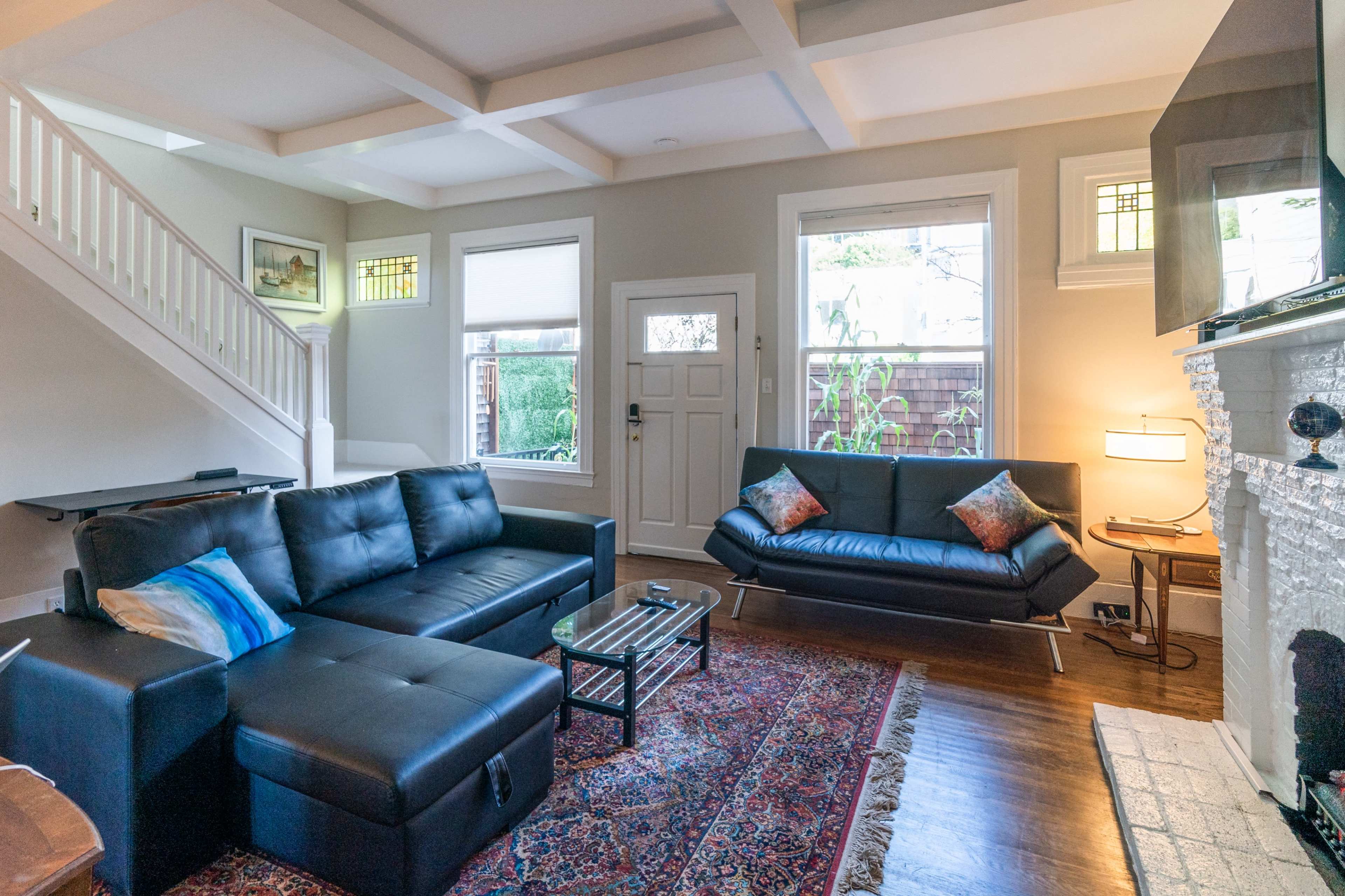 The living room features a black leather sectional sofa, a glass coffee table, and a light-colored stone fireplace, illuminated by natural light from the windows.