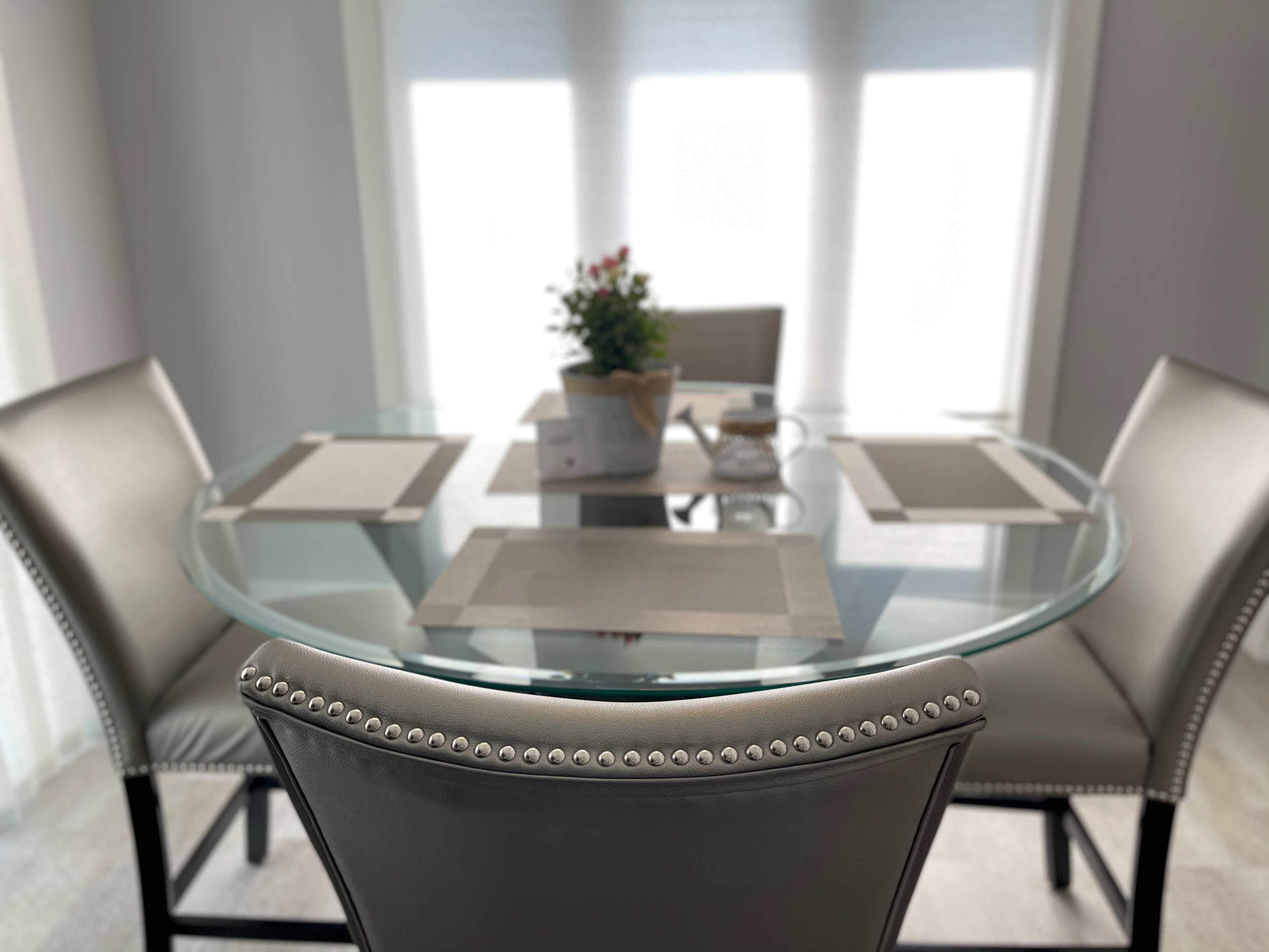 A round glass dining table is set with placemats and surrounded by upholstered chairs in a well-lit room.