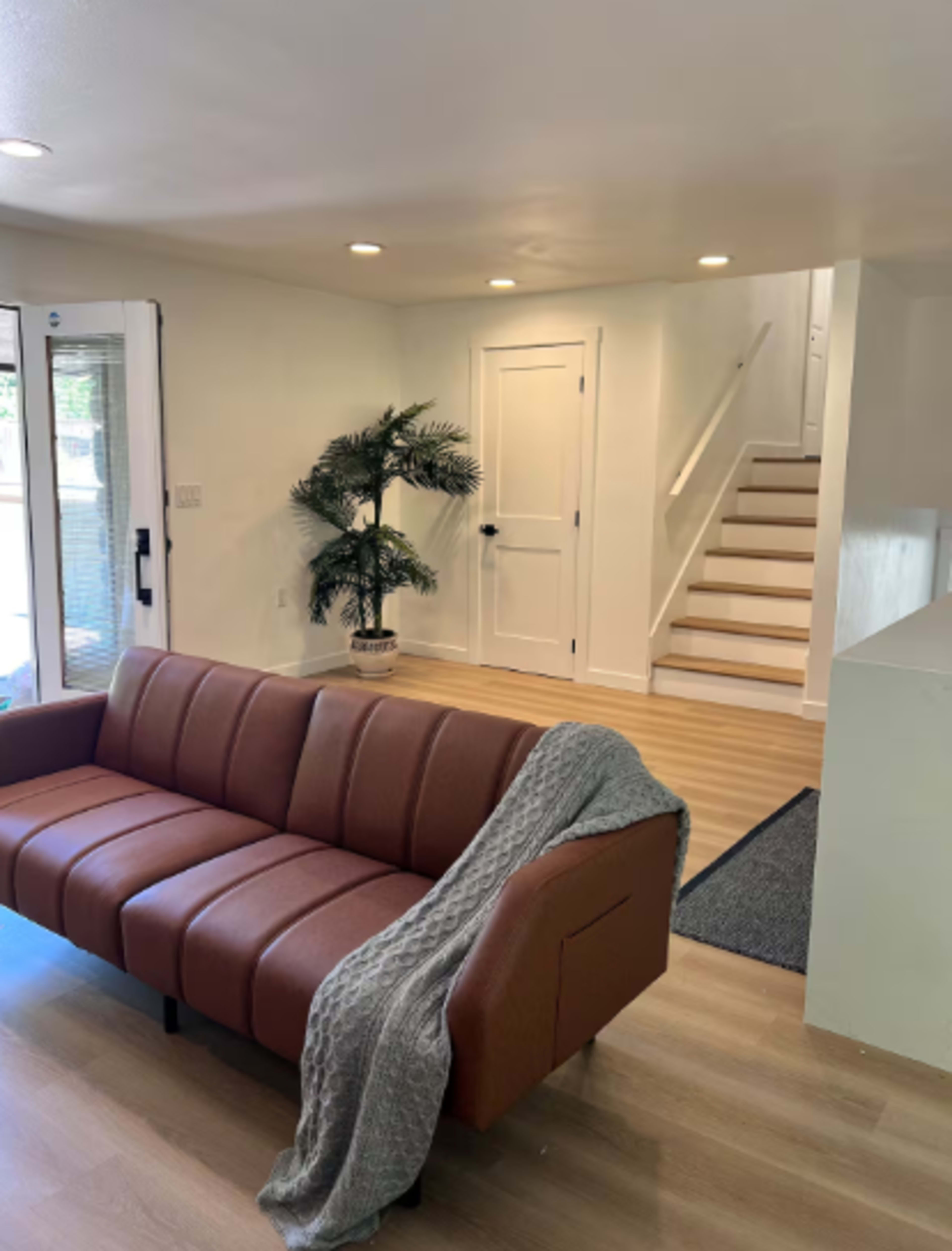 The image shows a modern living area featuring a brown leather sofa, a potted plant, and a staircase leading to an upper level.