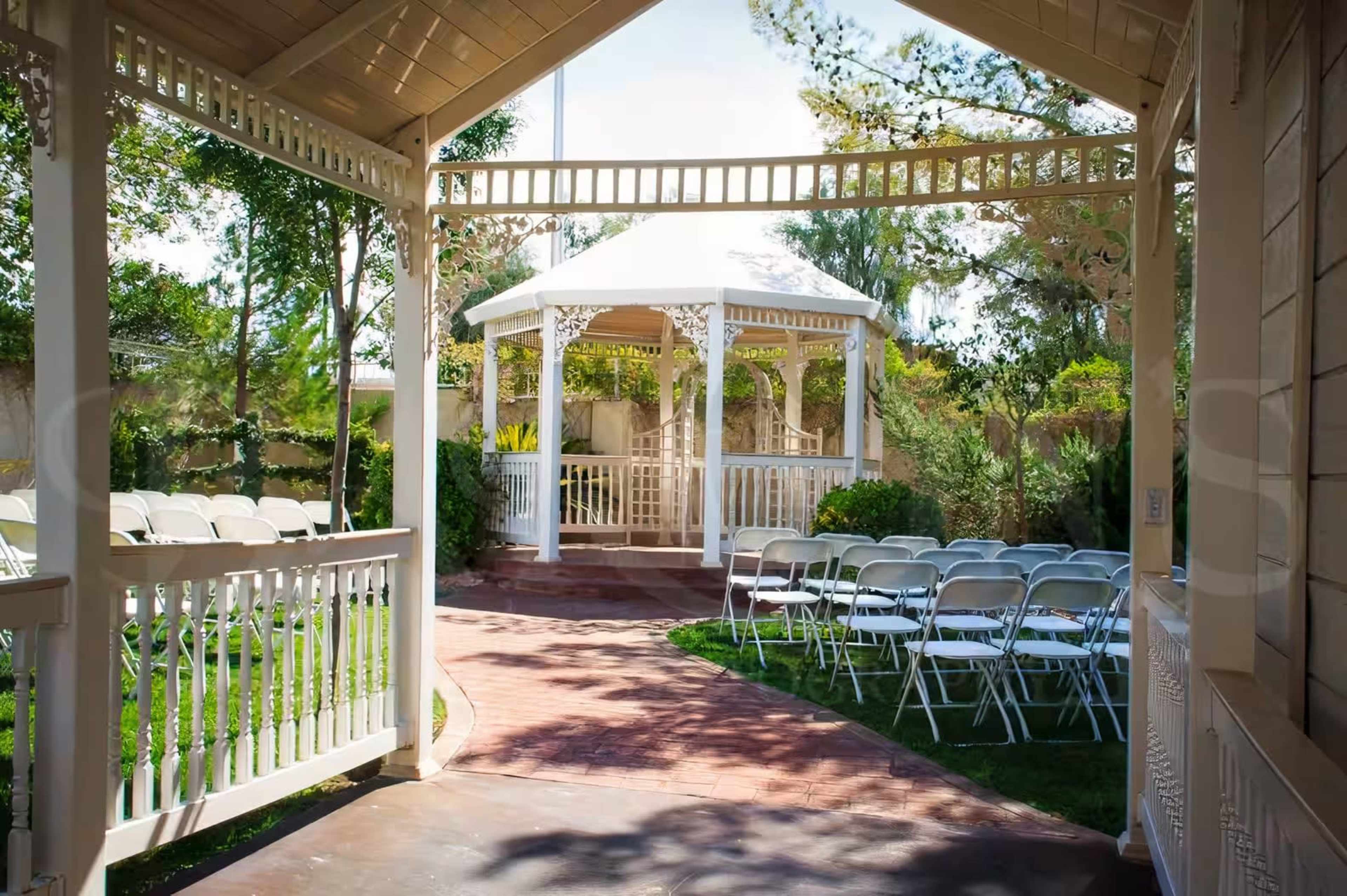 The image shows a gazebo in a landscaped garden with white folding chairs arranged in front for an event.