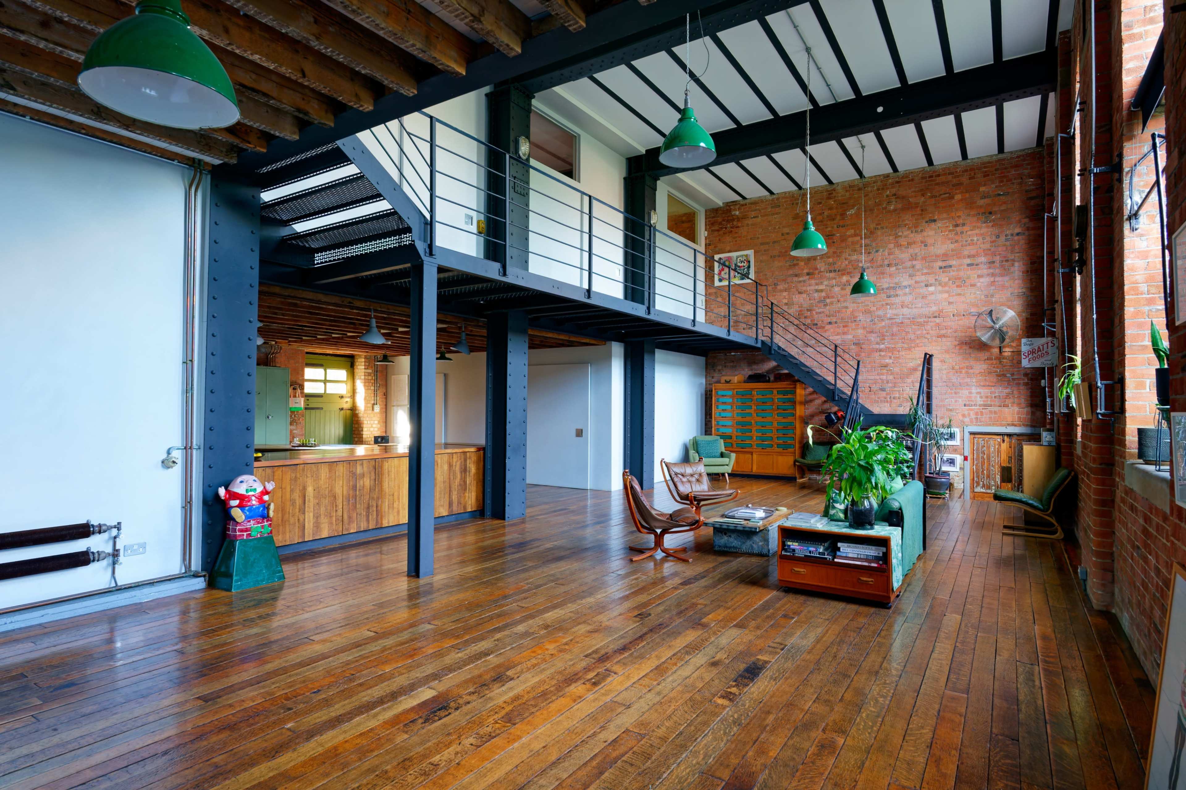 The image depicts a spacious industrial-style loft with exposed brick walls, wooden flooring, and a staircase leading to an upper level.