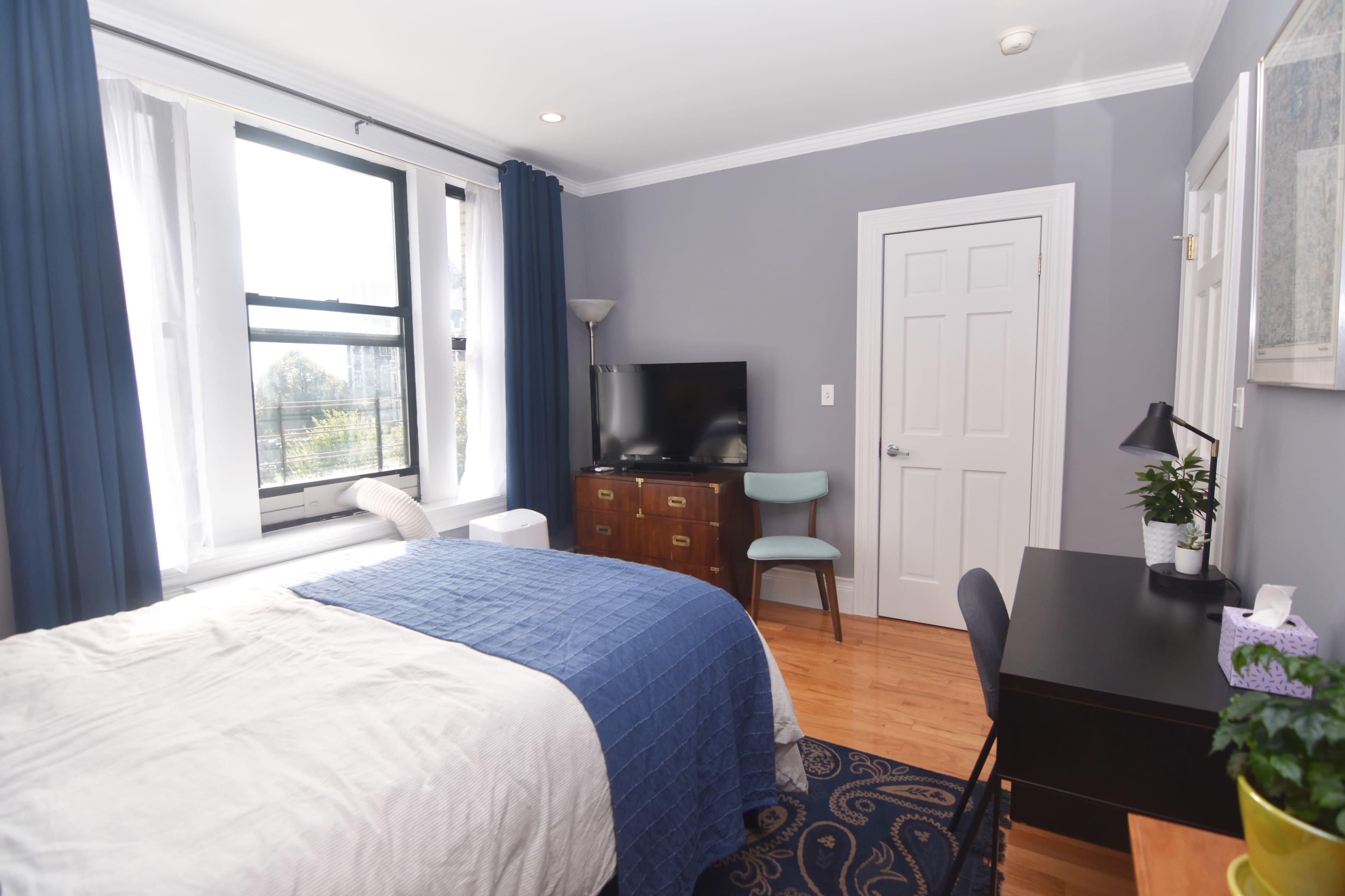 The image shows a simple bedroom with a bed, a desk and chair, a television, and a window with curtains.