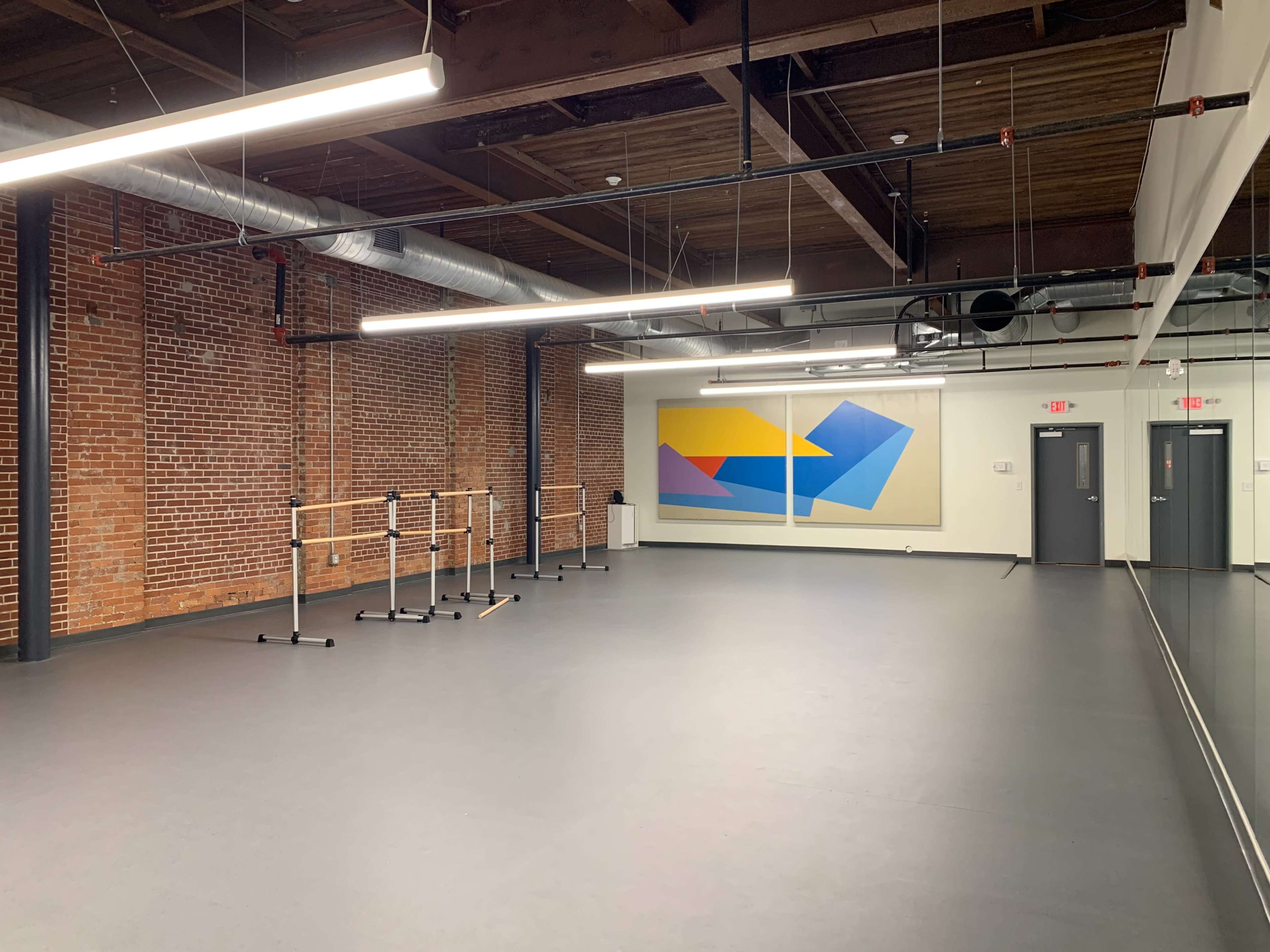 A spacious dance studio with a black floor, exposed brick walls, large mirrors, and a colorful abstract mural on one wall.