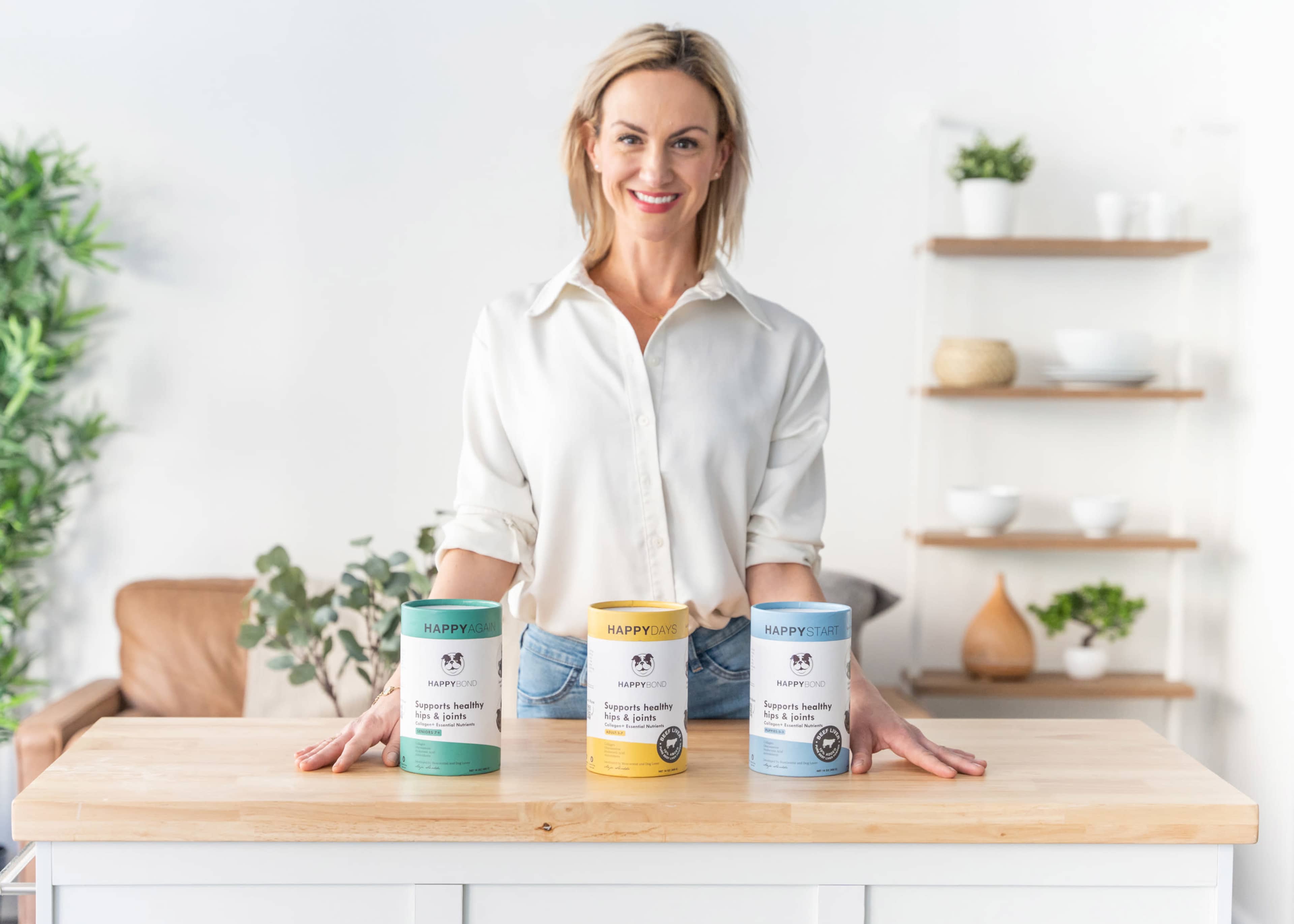 A person stands behind a wooden table displaying three containers of pet supplements in a bright, modern kitchen setting.