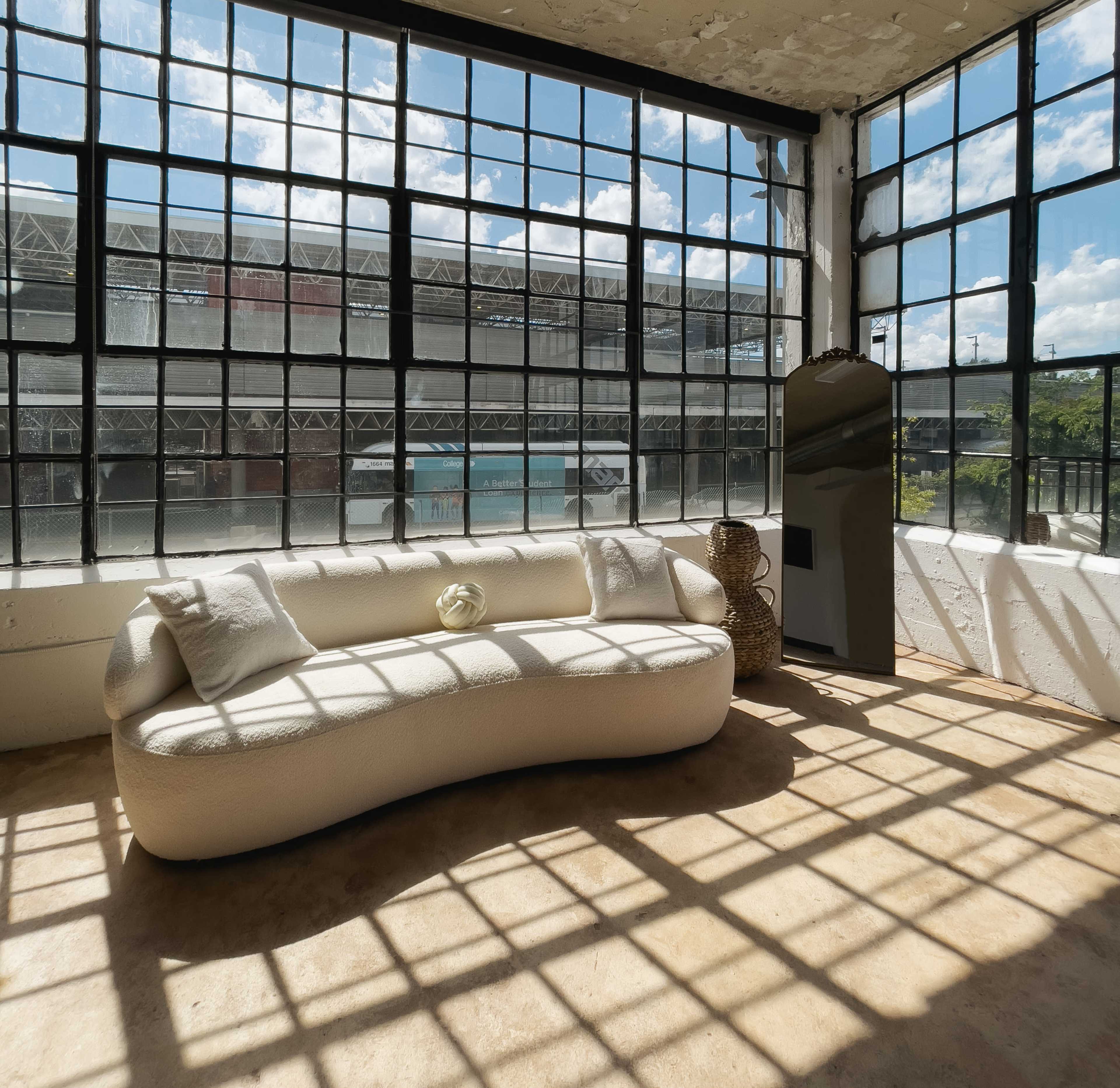 A curved white sofa is positioned in front of large industrial windows filled with natural light, revealing an outdoor view of a structure.
