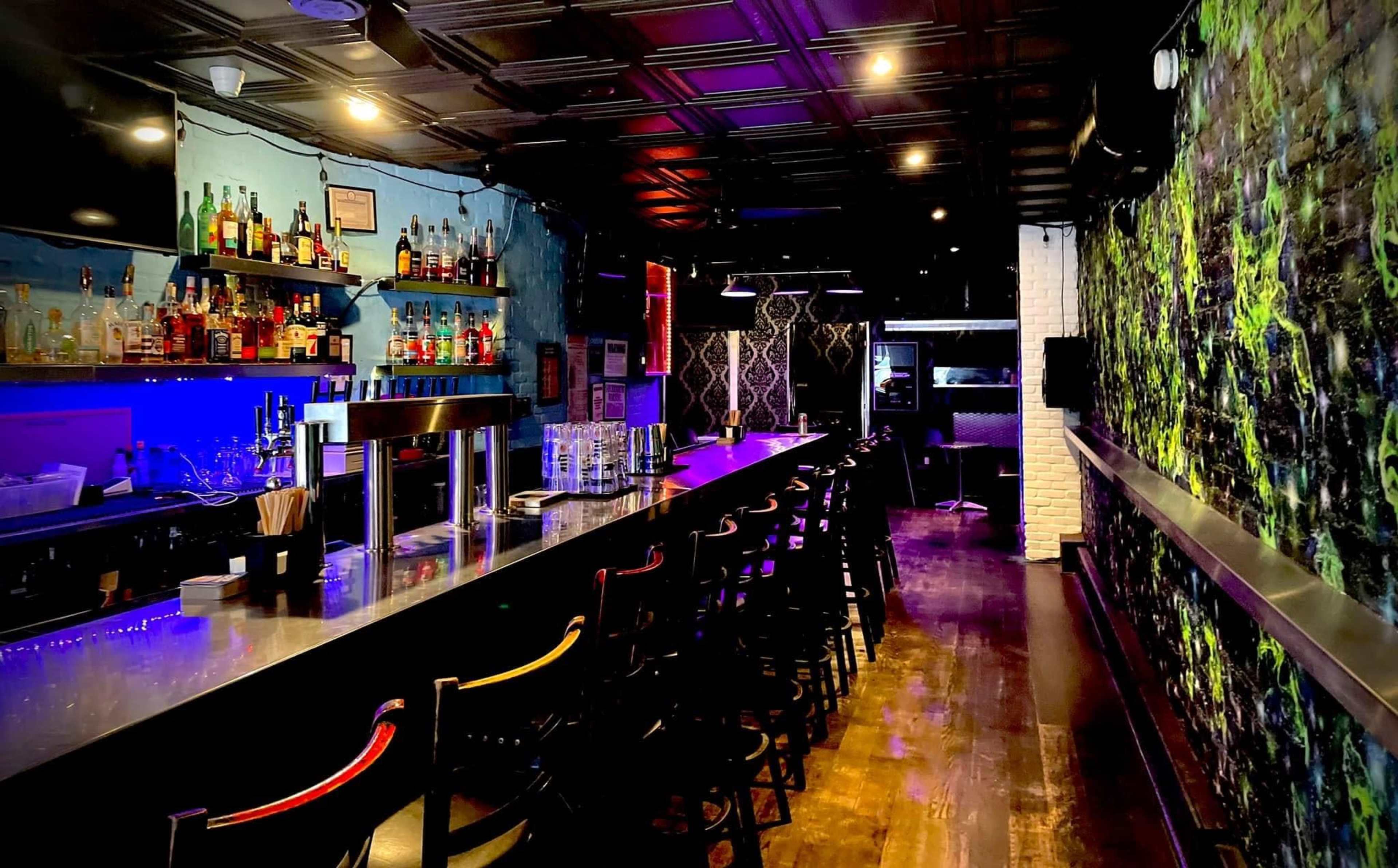 The image shows a modern bar with a long countertop, shelves lined with various liquor bottles, and colorful lighting effects.